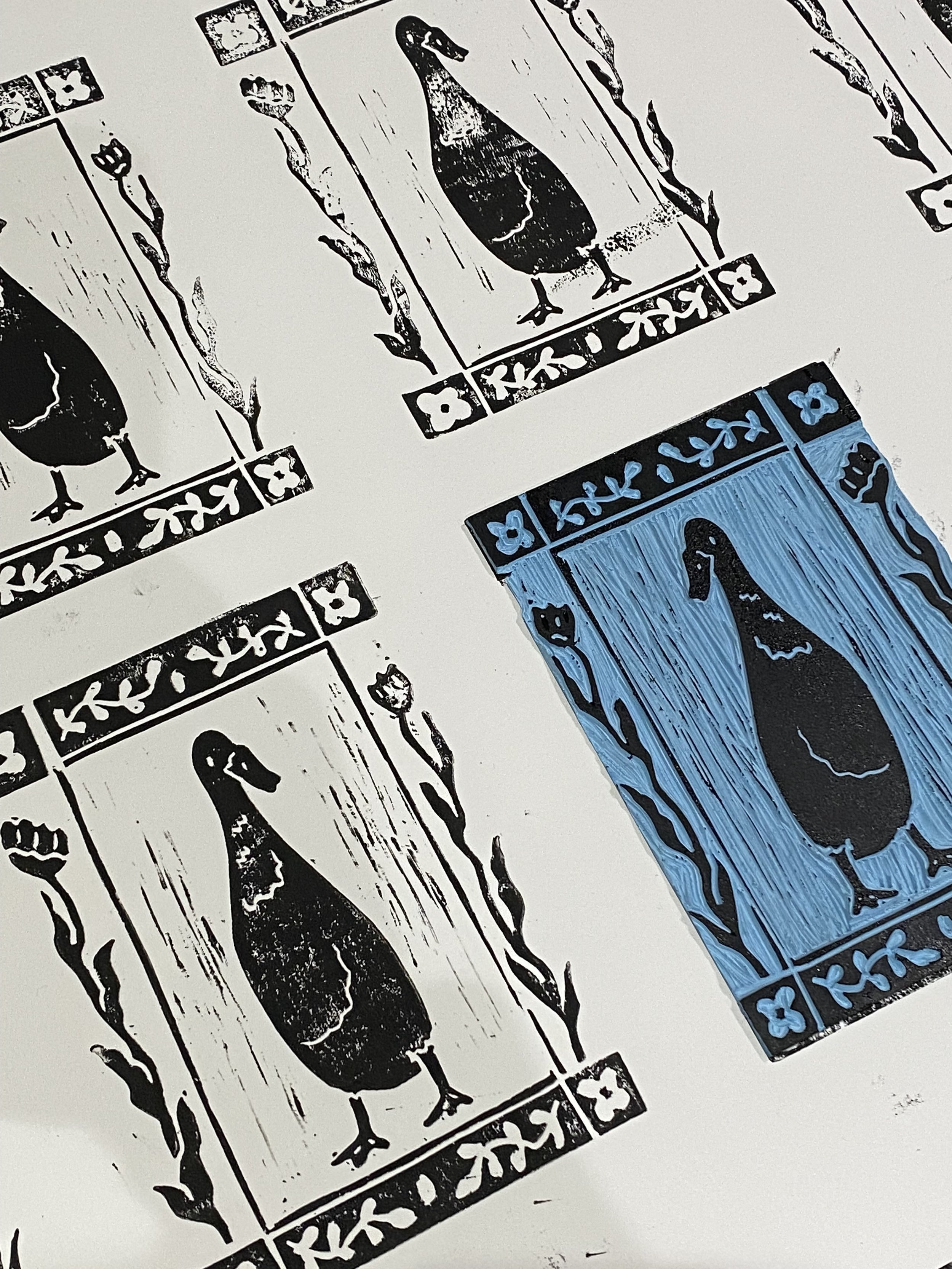 A black lino printed illustration of a goose on a piece of paper, with decorative floral borders.