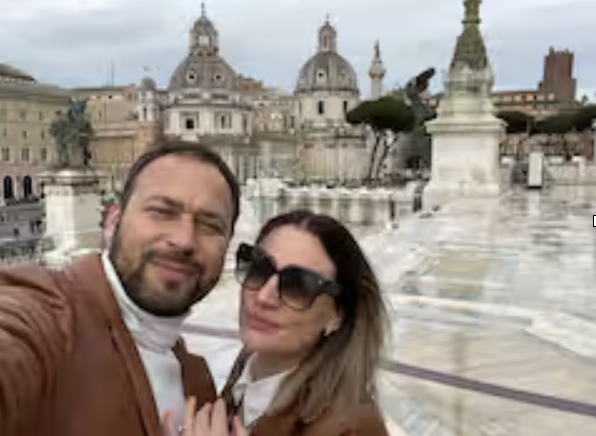 A man and woman taking a selfie on a rooftop with historic buildings, domed structures, and statues in the background.