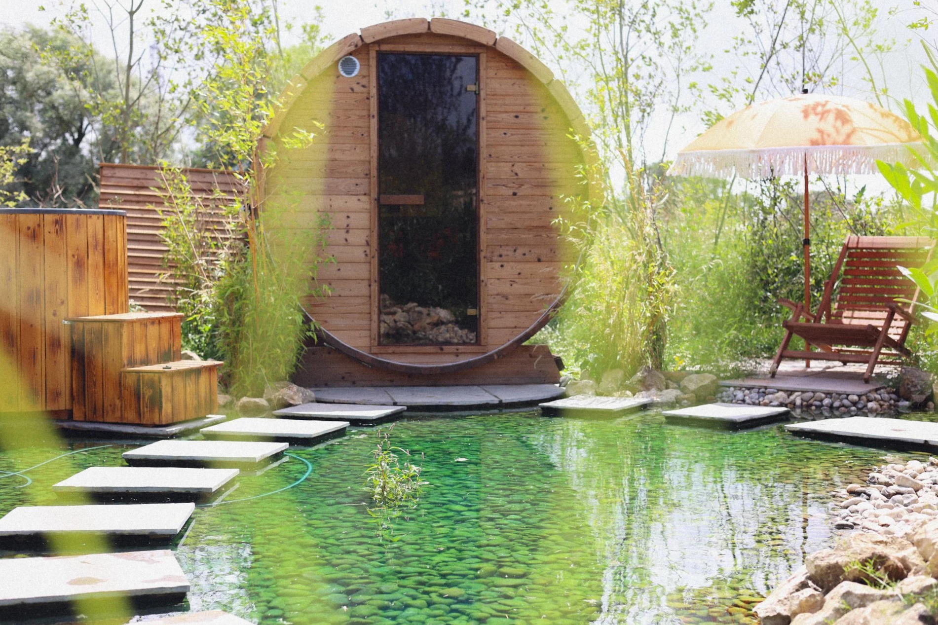 A small round wooden sauna beside a pond with stepping stones, a wooden bench with an umbrella, and greenery in the background.