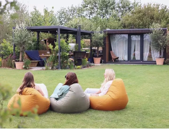 Three women sitting on bean bags on a grassy lawn, engaged in conversation, with a modern backyard patio and garden area in the background.