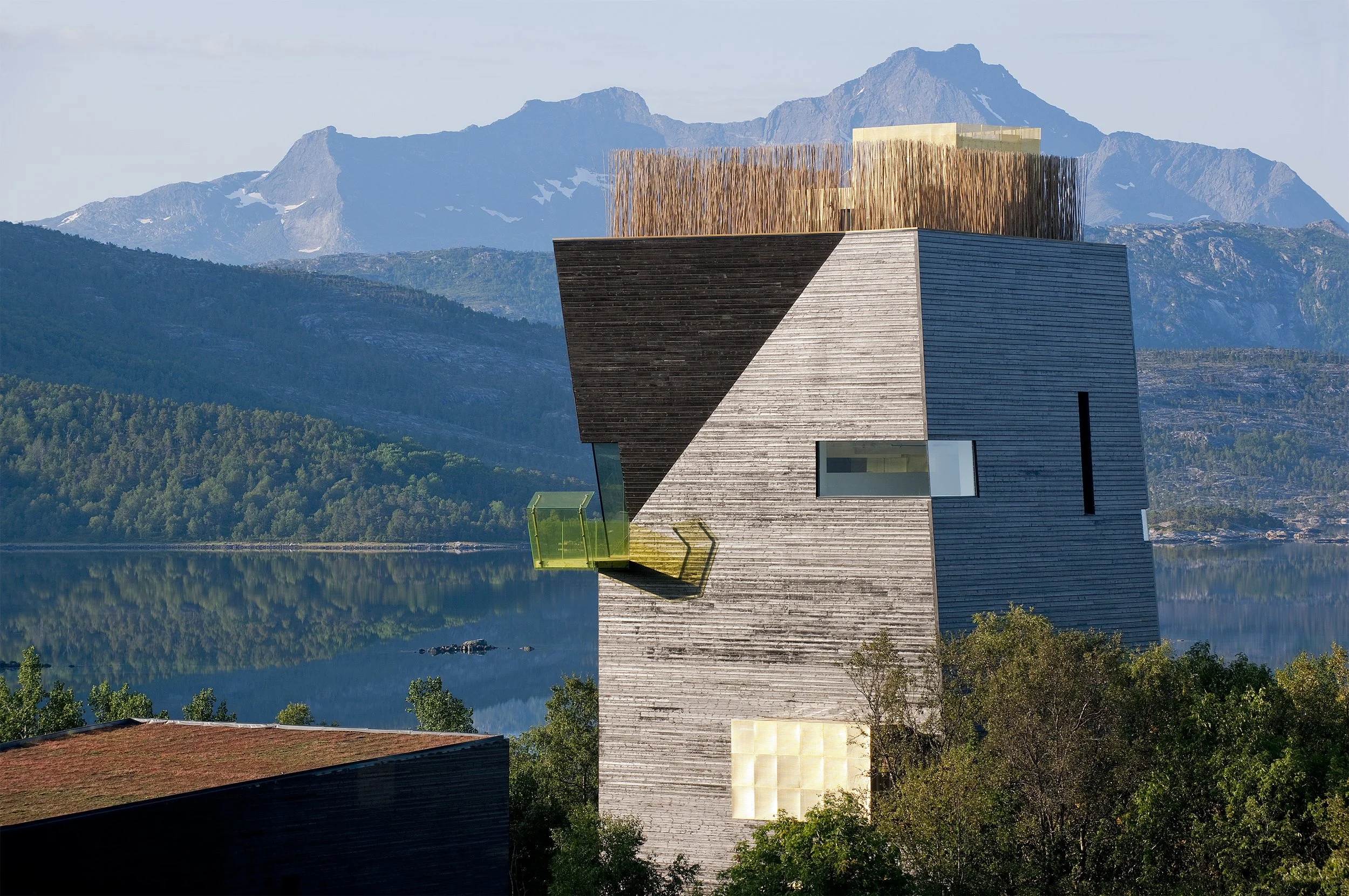 En moderne bygning med geometriske former, store vinduer og en utendørs terrasse, omgitt av natur med vann og fjell i bakgrunnen.