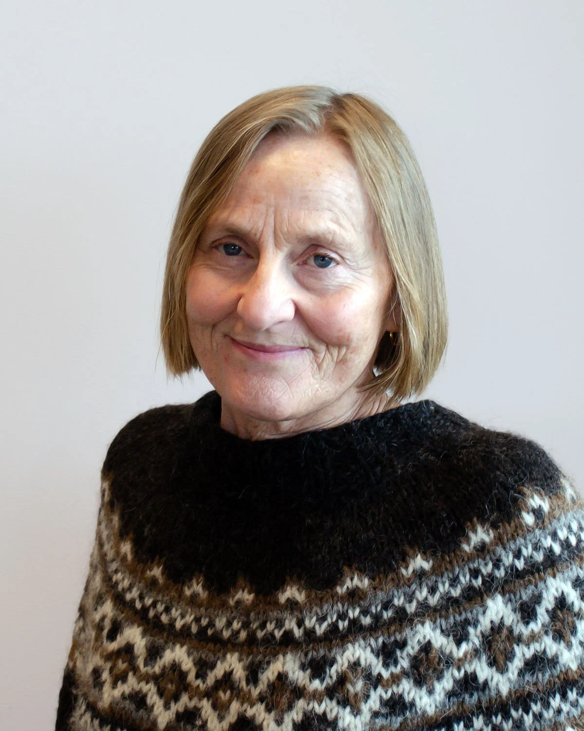 Portrett av en eldre kvinne med kort, blond hår, smilende, iført en svart og brunsvart mønstret strikkegenser, stående foran en hvit vegg.
