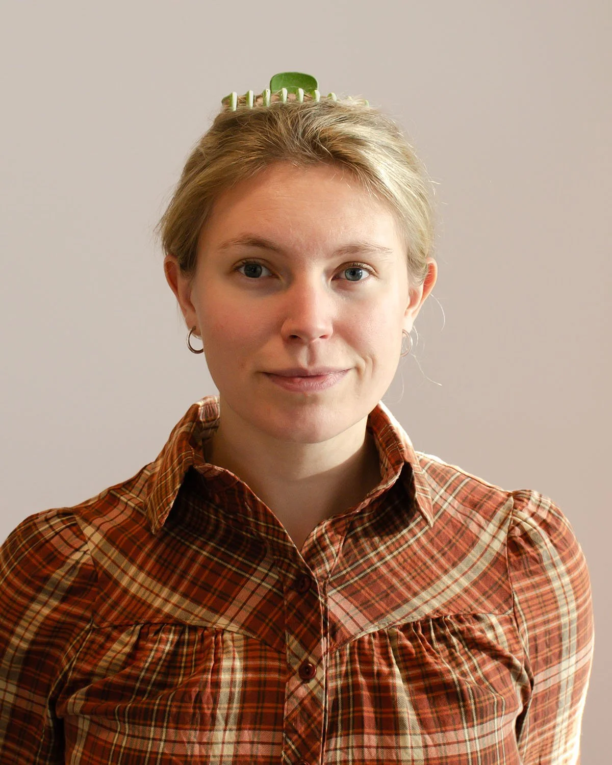Kvinne med blonde kort hår og blå øyne, smilende, iført rutete skjorte, med hårklipp og øredobber, står mot en nøytral bakgrunn.