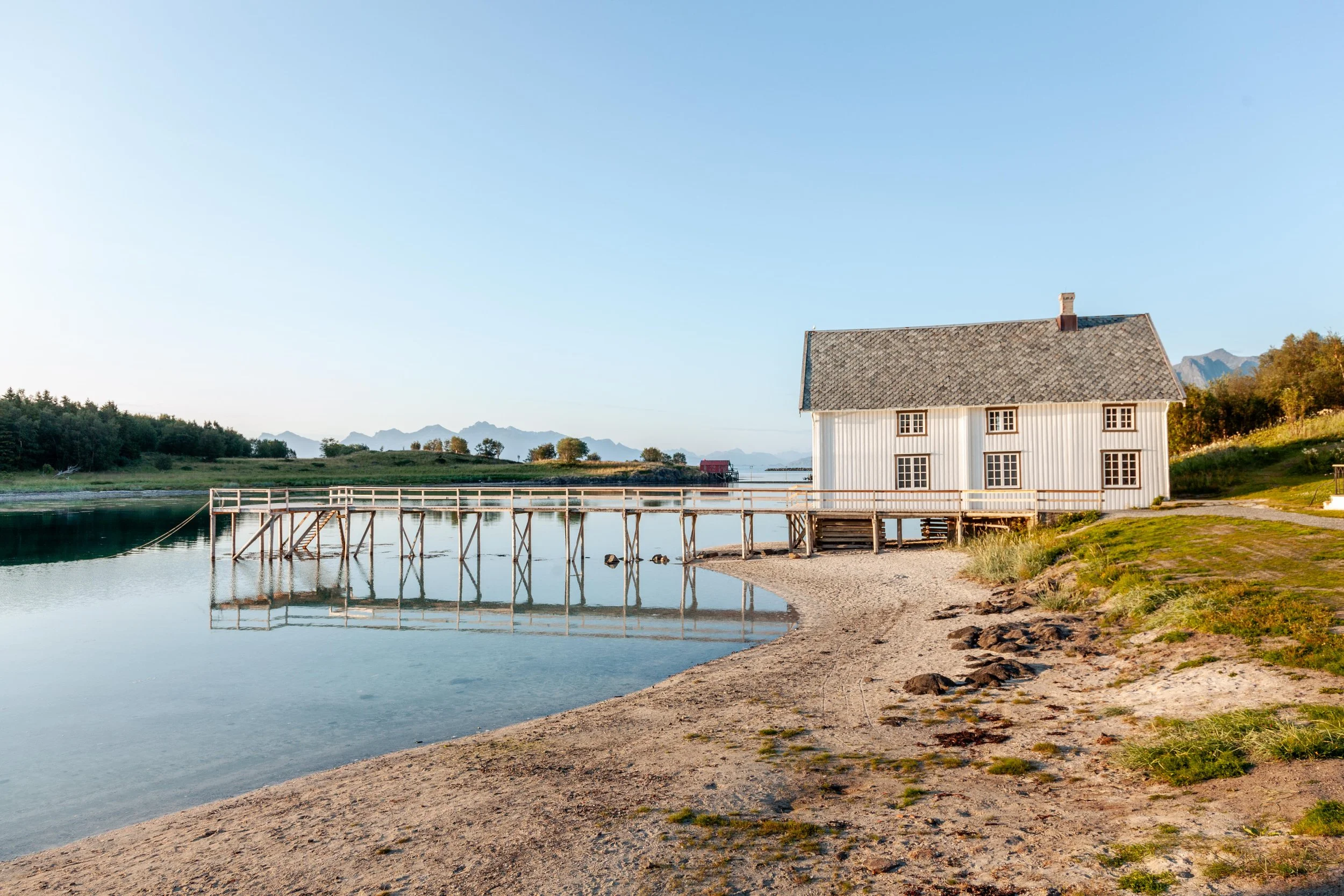 En hvit hus ved vann med trebro og sandstrand, omgitt av grønne åser og fjell i bakgrunnen, under en klar blå himmel.