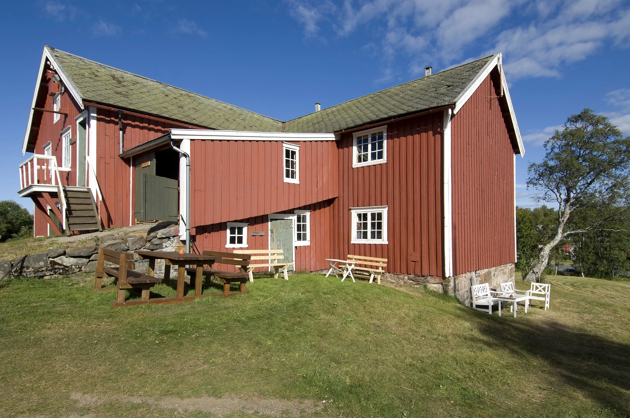 En rød tradisjonell svensk stabskur, med hvite vindusrammer og trapper, som står på en grønn gresslette under en blå himmel med noen skyer.