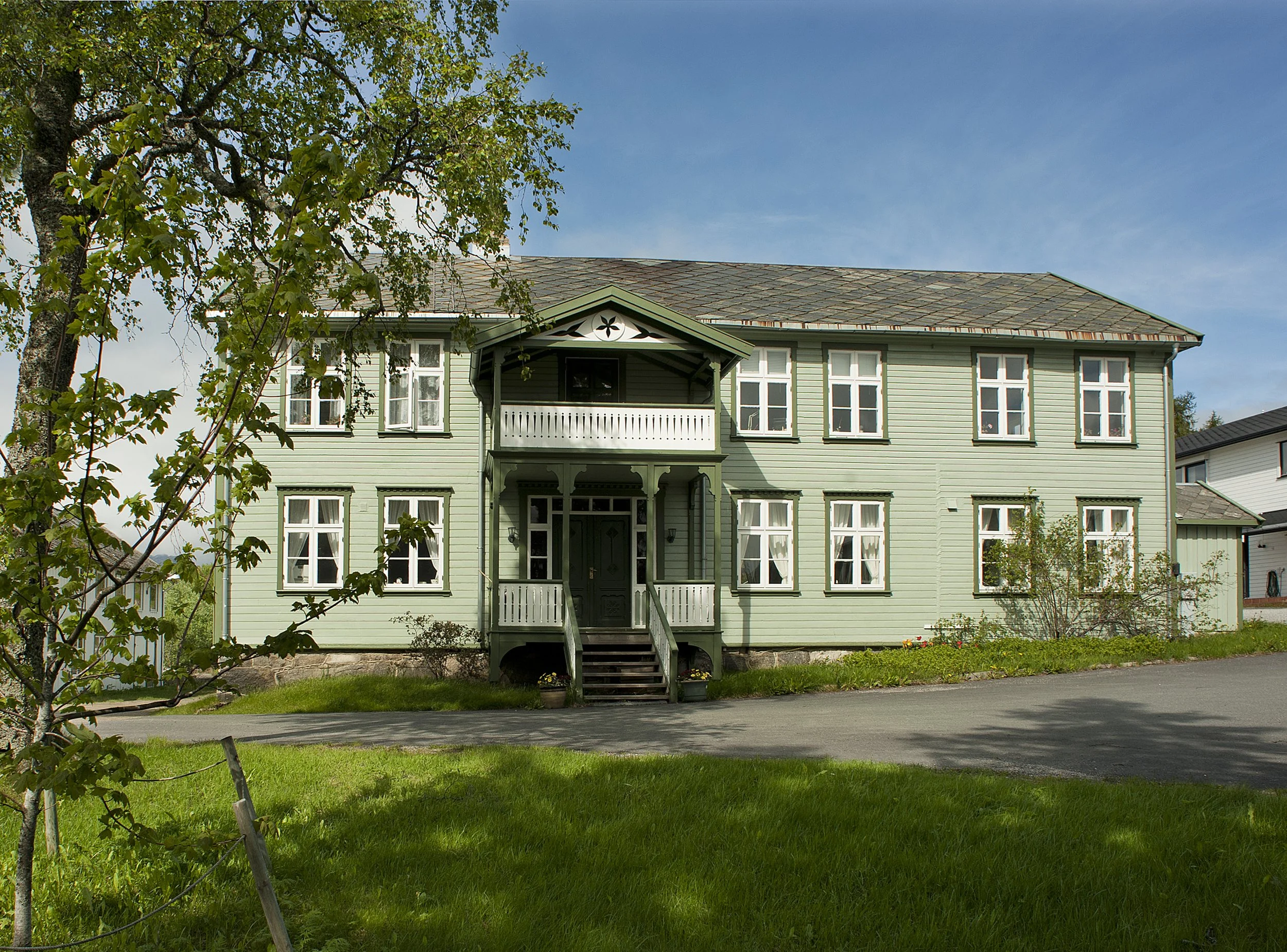 Et lys grønt hus med veranda og mange vinduer, omgitt av grønne planter og trær, under en klar blå himmel.
