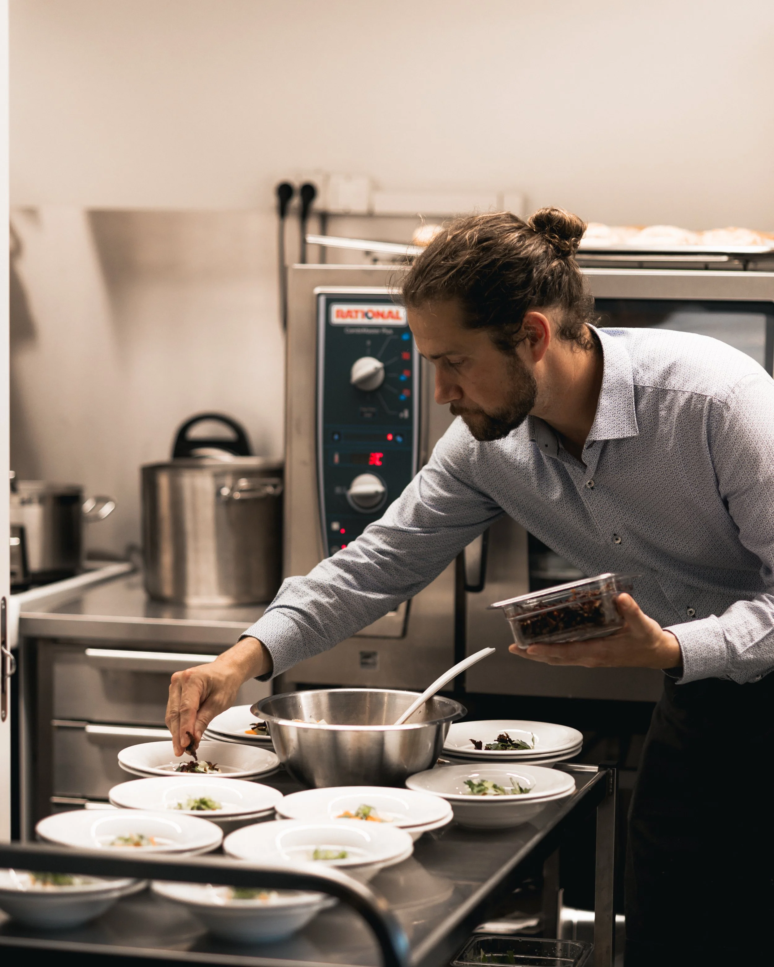 En mann som serverer mat i et profesjonelt kjøkken, med flere tallerkener av ferdige retter på bordet.