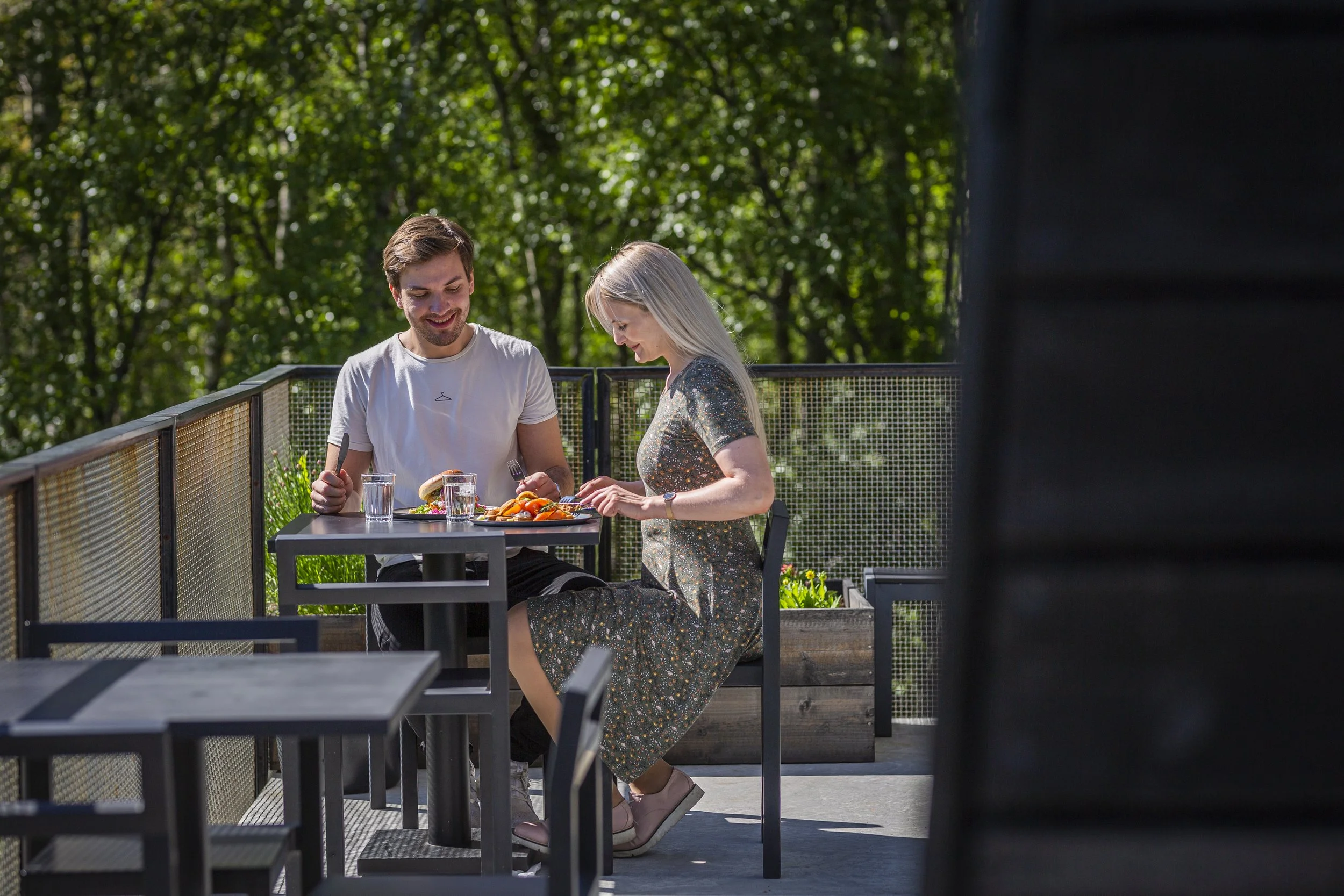 En mann og en kvinne sitter ved et bord på en utendørs terrasse omgitt av grønne trær, de deler et måltid og smiler.