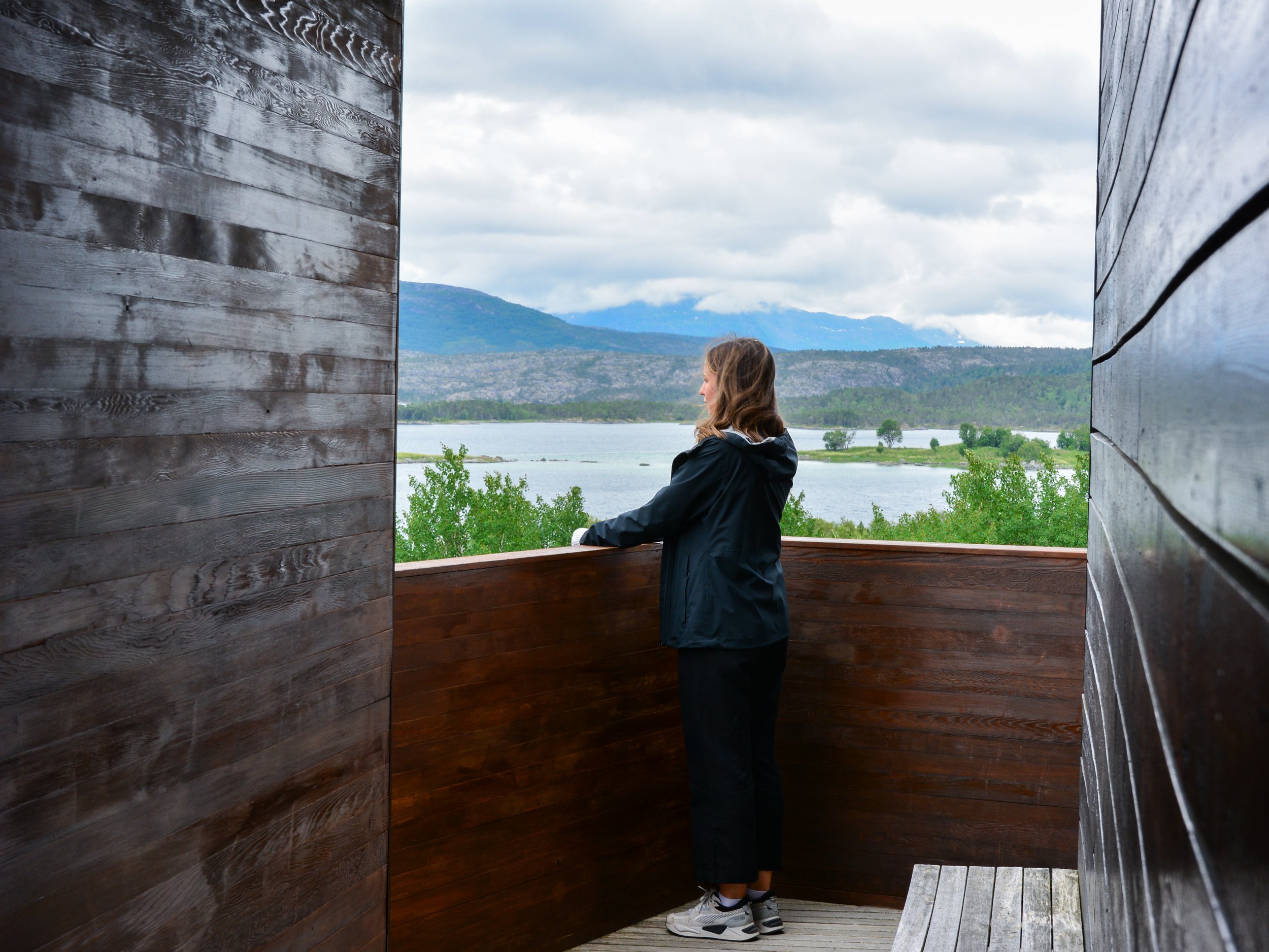 En kvinne står på en balkong med utsikt over en innsjø og fjell i bakgrunnen, omgitt av trevegger.