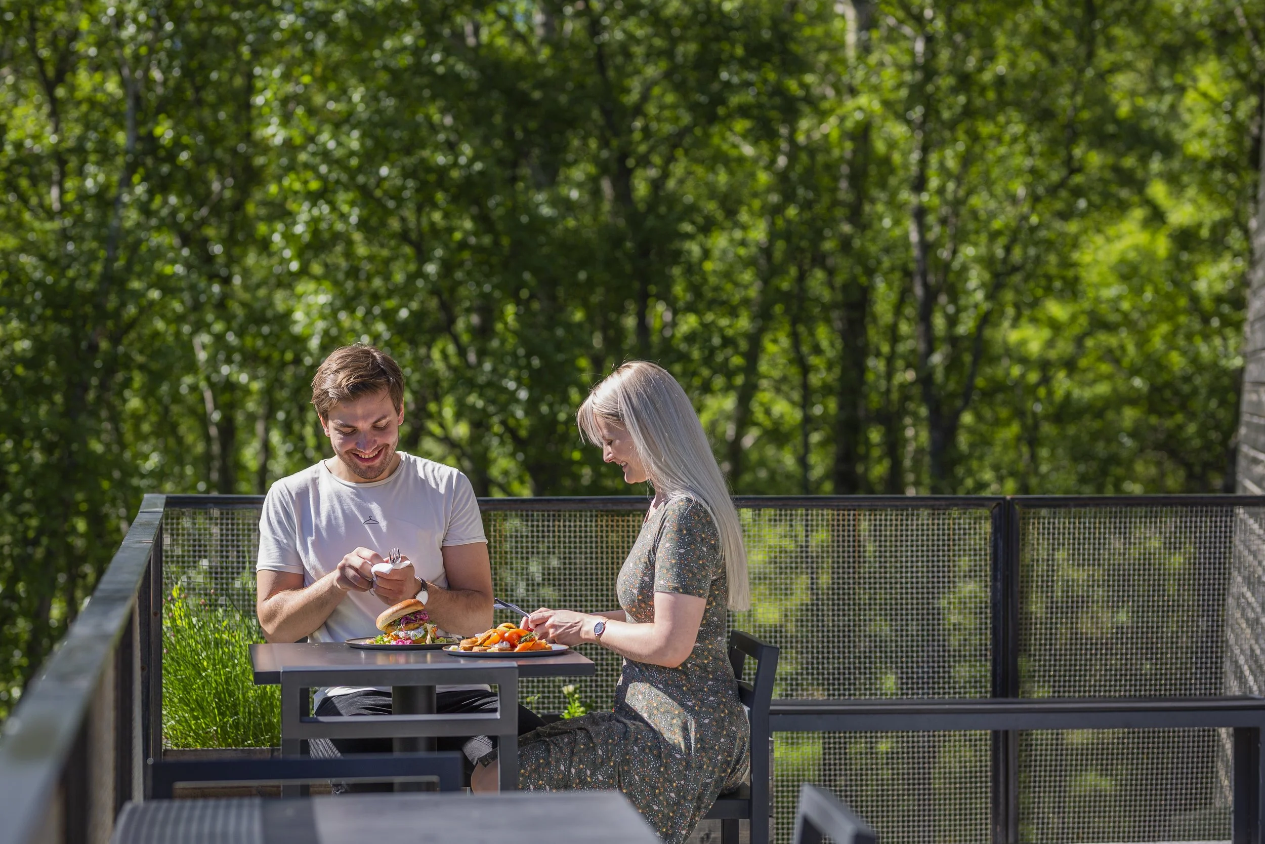 To personer som spiser ute i en park med grønt løvverk. De smiler og deler mat på en utendørs restaurant eller kafé.