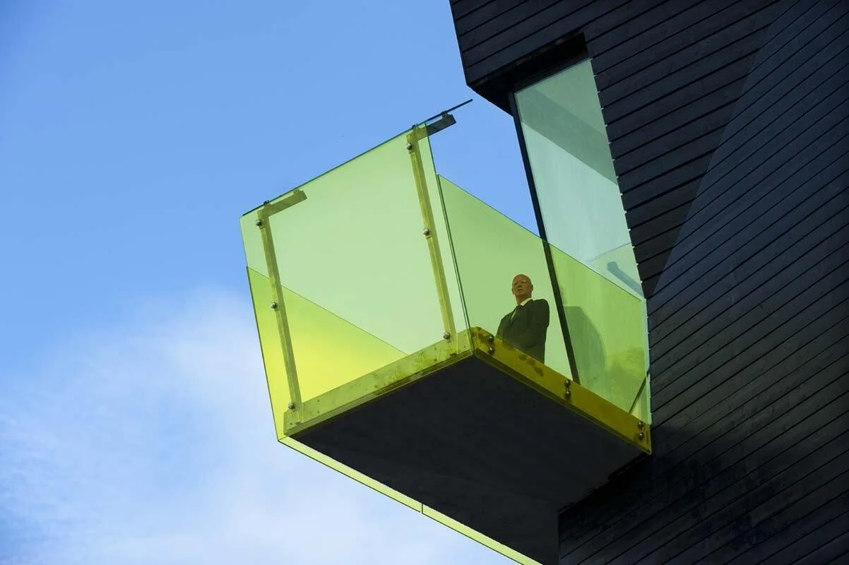 En mann i dress står på en gulgrønn glassballkong på et moderne bygg mot en blå himmel.