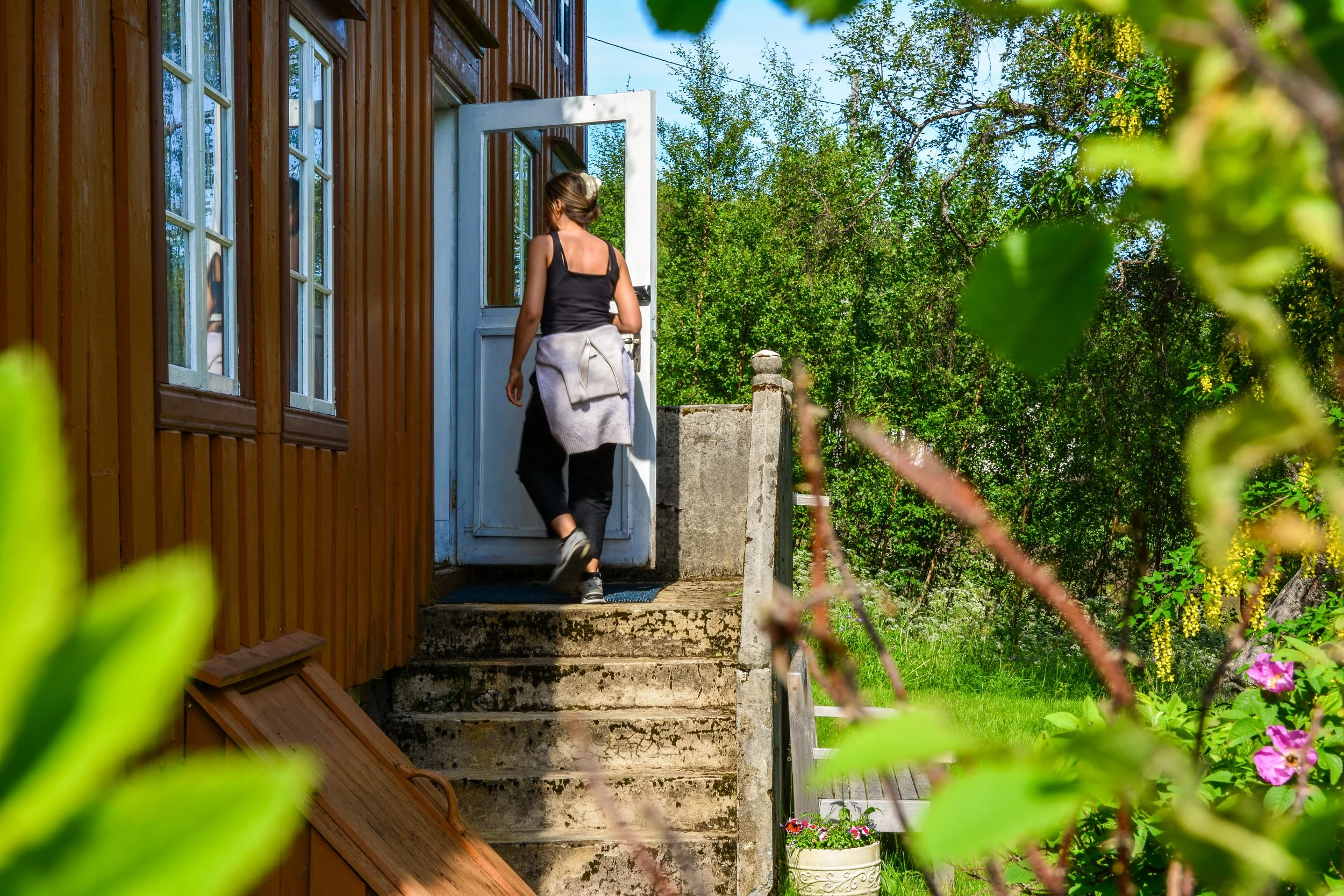 En kvinne går inn gjennom en hvit dør på terrassen til en trebrygger i en grønn hage med blomster.