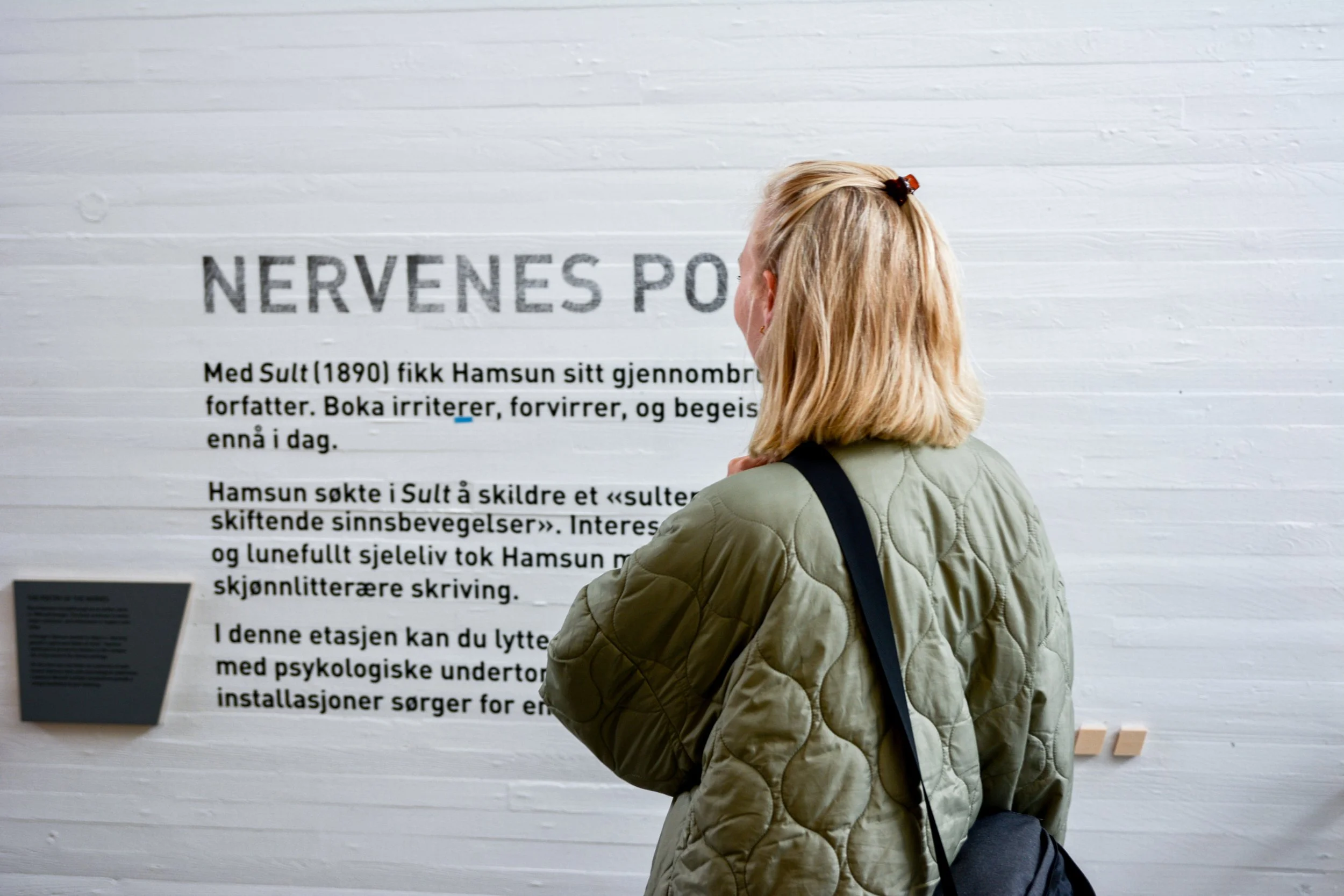 En kvinne med blondt hår, iført grønn jakke og svart veske, står foran en utstilling eller informasjonstavle på en hvit vegg.
