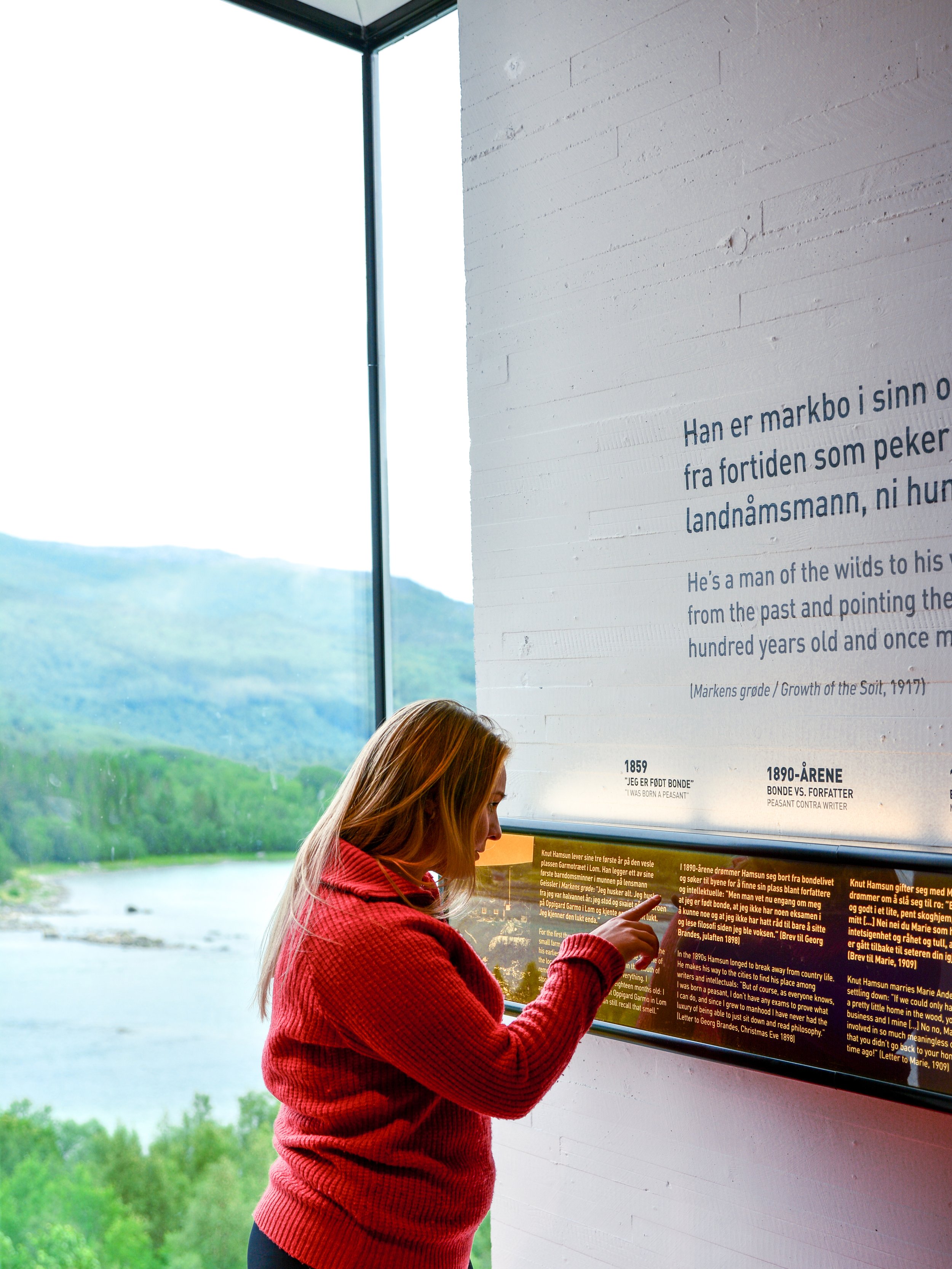 En kvinne med rød genser som ser på informasjonstavle inne med store vinduer og utsikt over grønne åser og innsjø ute.