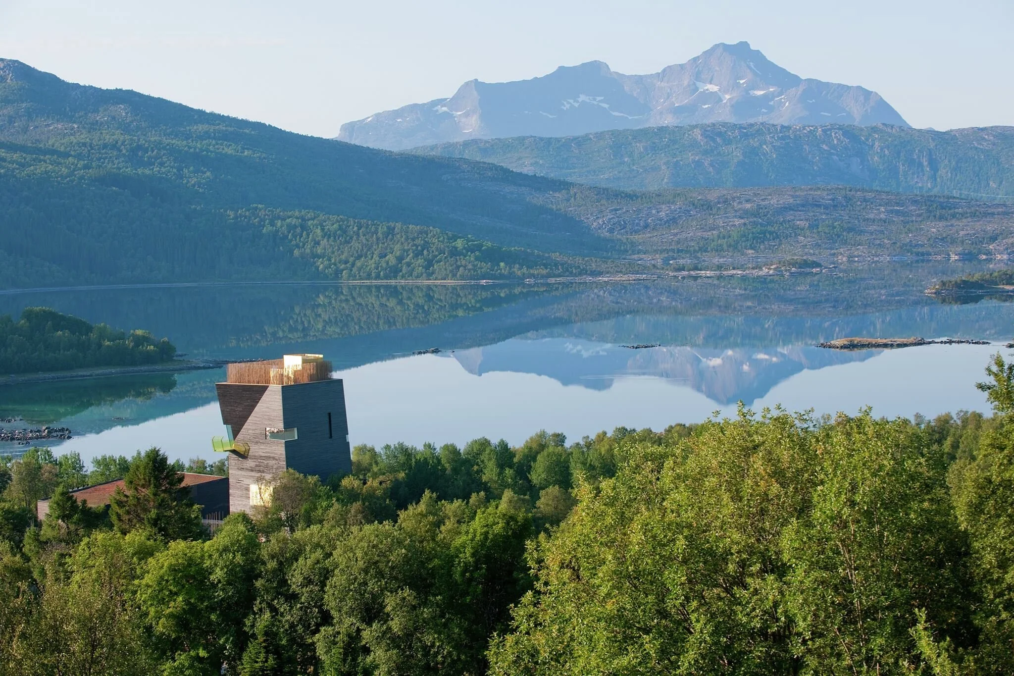 Fjellandskap med innsjø og moderne bygning i forgrunn, grønne trær, høye fjell i bakgrunnen, tydelig refleksjon av fjellene i vannet