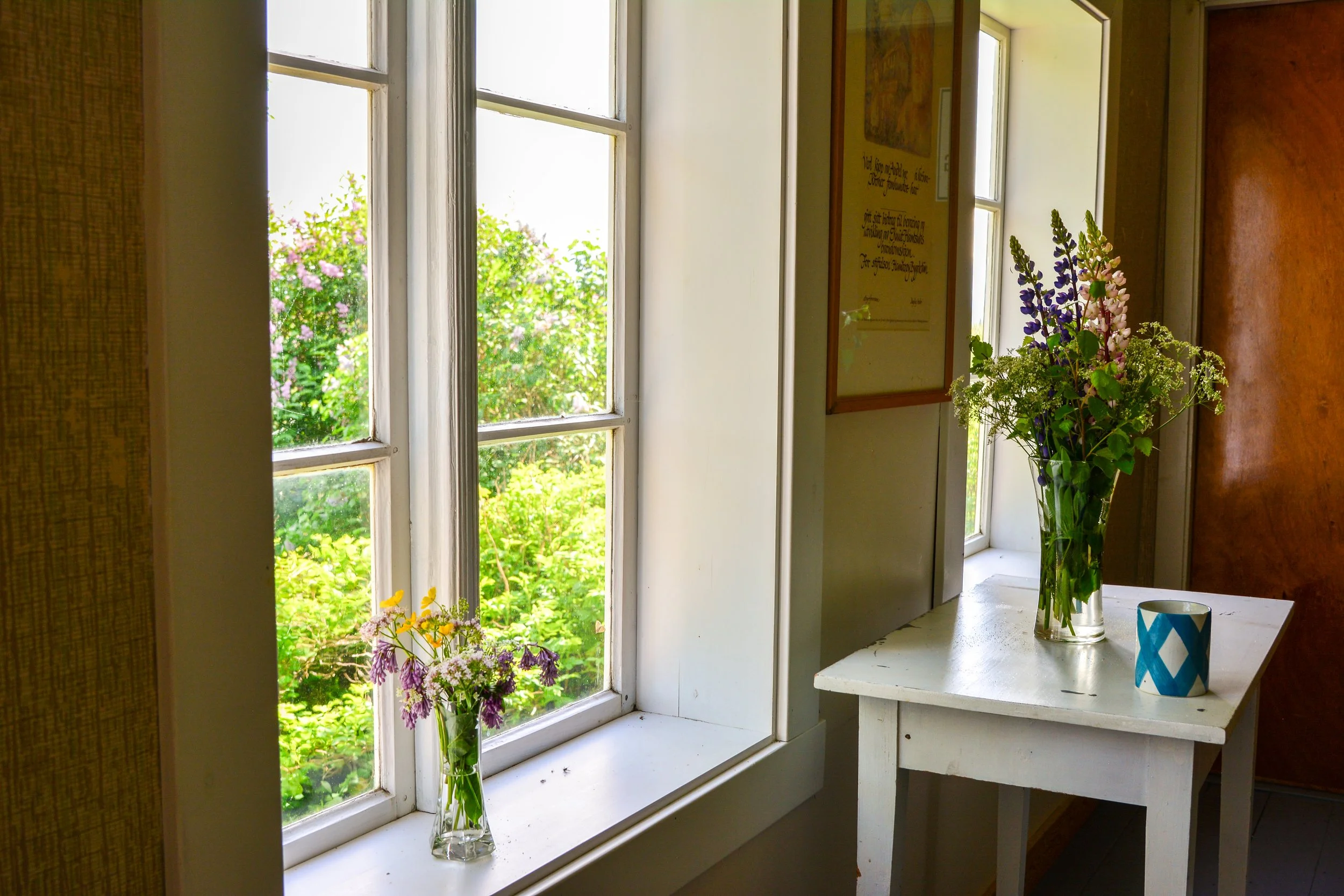 Vase med blomster på et vindusbord ved et vindu som viser grønne trær utenfor.