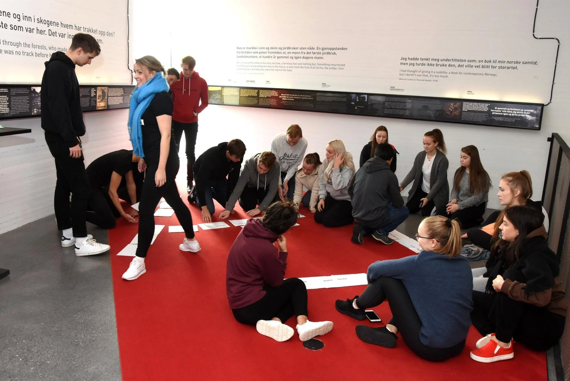 En gruppe unge mennesker samler seg i en gruppe på en rød matte med informasjonstavler på veggene, i et innendørs museum eller utstilling.