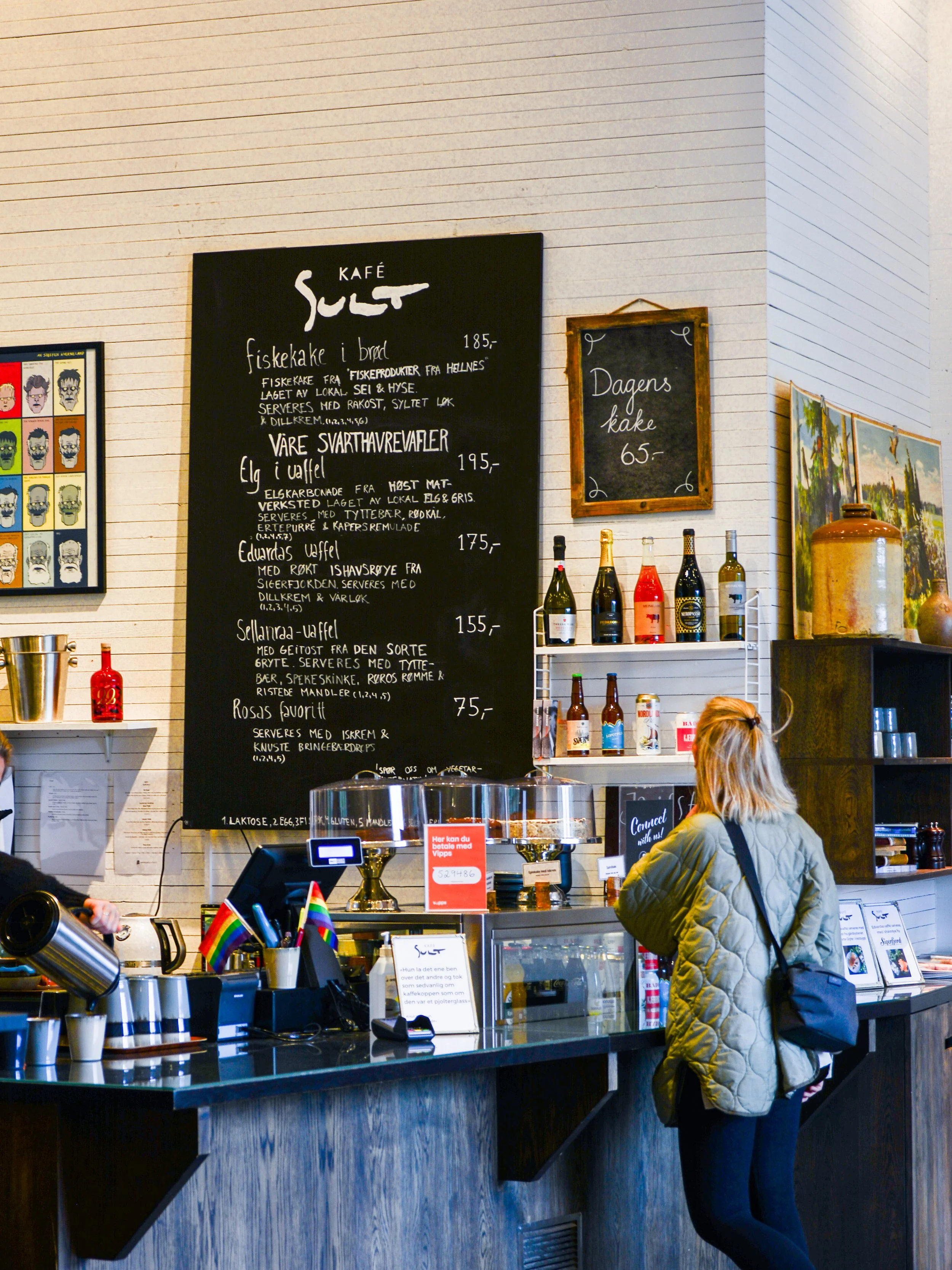 En kvinne med en grønn jakke og svart veske står foran en kaffebar med menyer skrevet på tavler på veggen. Det er flere flasker vin og andre drikkevarer på hyllen bak disken.