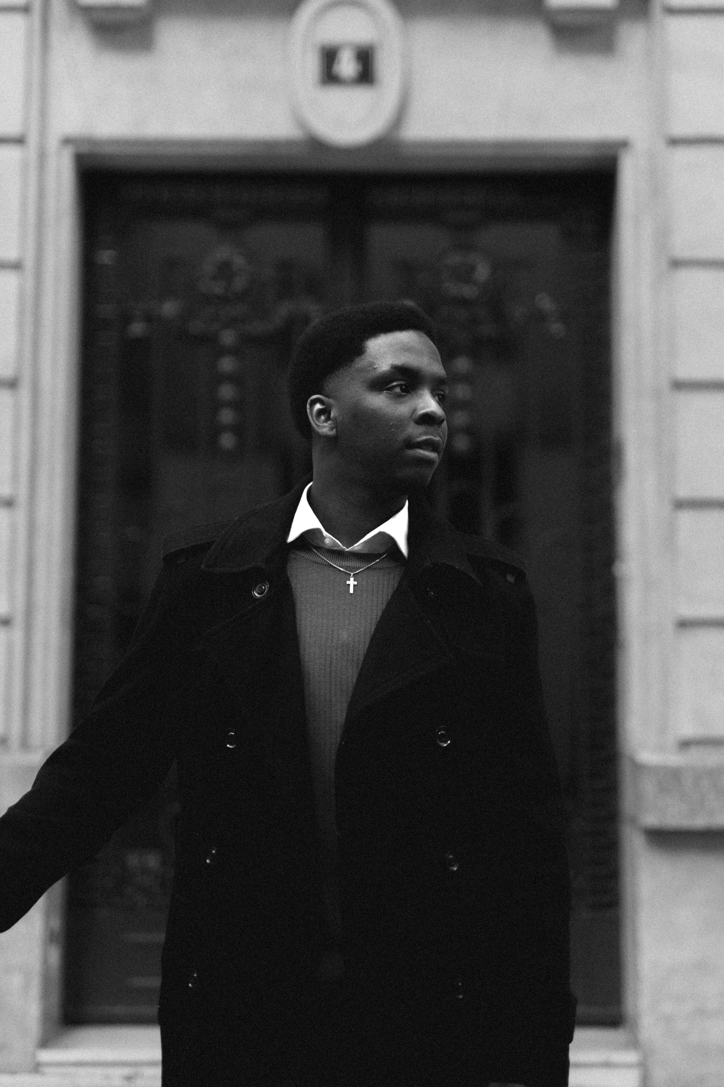 Jeune homme noir en manteau noir, portant une chemise blanche, un pull gris et une chaîne avec une croix, debout devant une porte ornée, en noir et blanc.