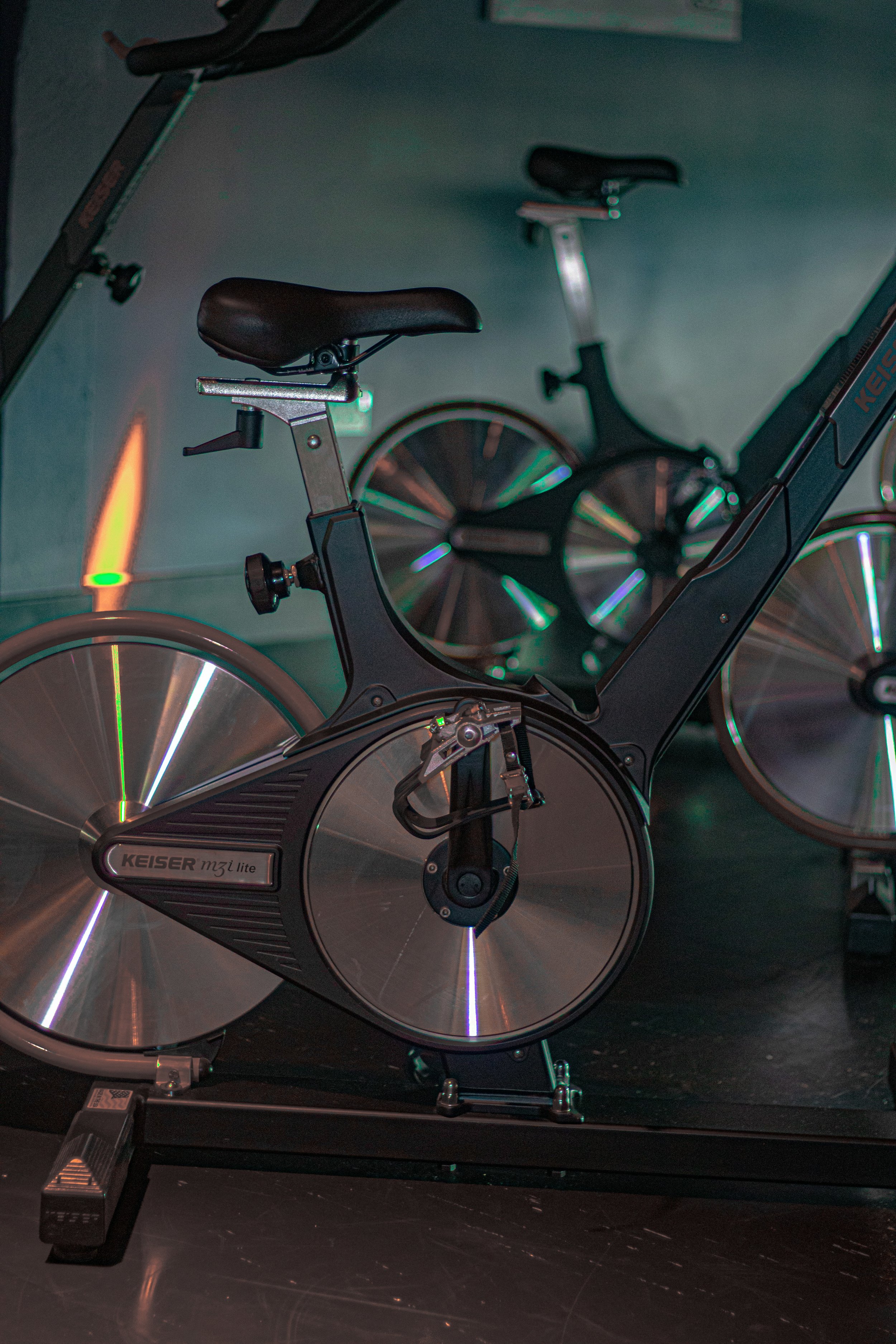 Electric exercise bike with a black frame and metallic flywheels, with a second similar bike in the background, in a dimly lit room.