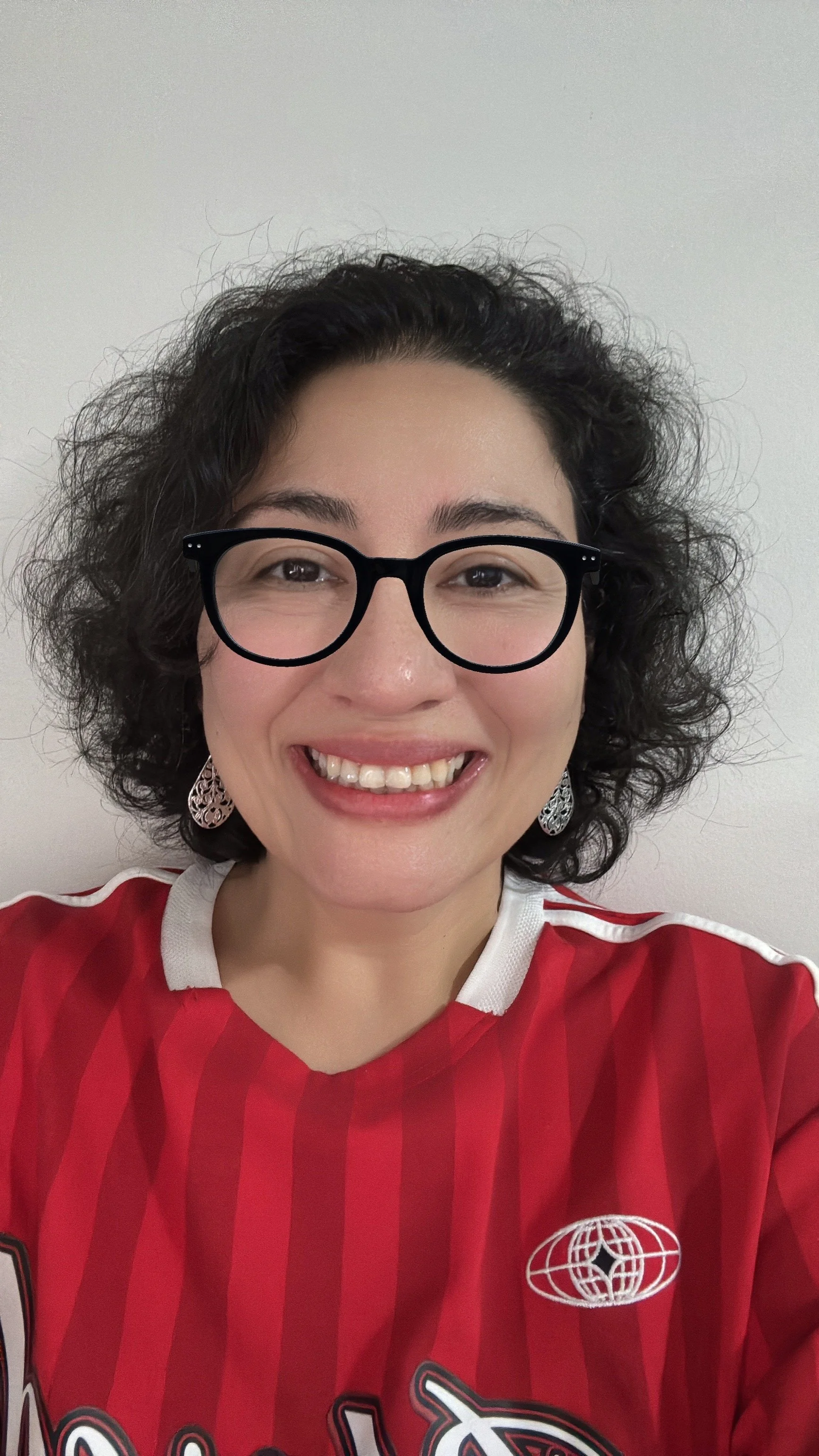 A woman with dark curly hair wearing glasses, earrings, and a red sports jersey smiling at the camera against a plain white background.