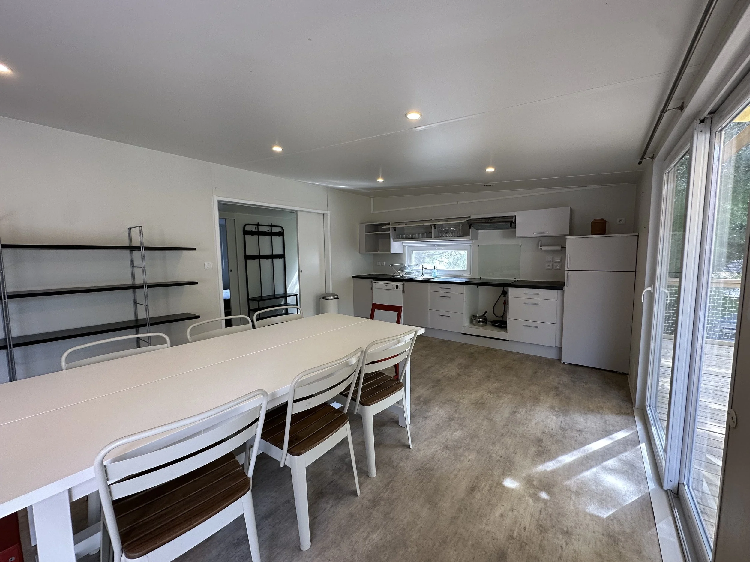 Cuisine spatieuse avec grande table blanche, chaises en bois et métal, cuisine blanche avec plan de travail noir, réfrigérateur blanc, et grande fenêtre en verre donnant sur un balcon en bois.