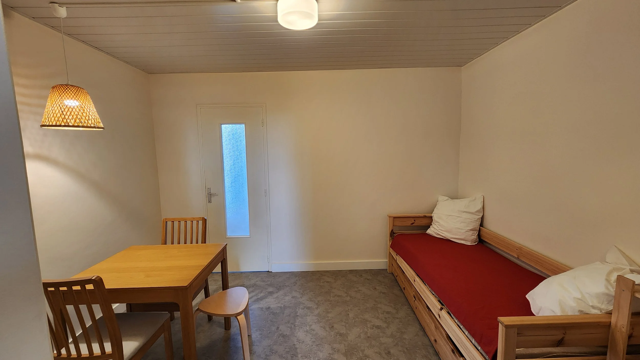 Pièce simple avec un lit en bois, une table et trois chaises en bois, deux oreillers sur le lit, une porte avec une vitre opaque, une suspension en rotin et un plafond en bois.