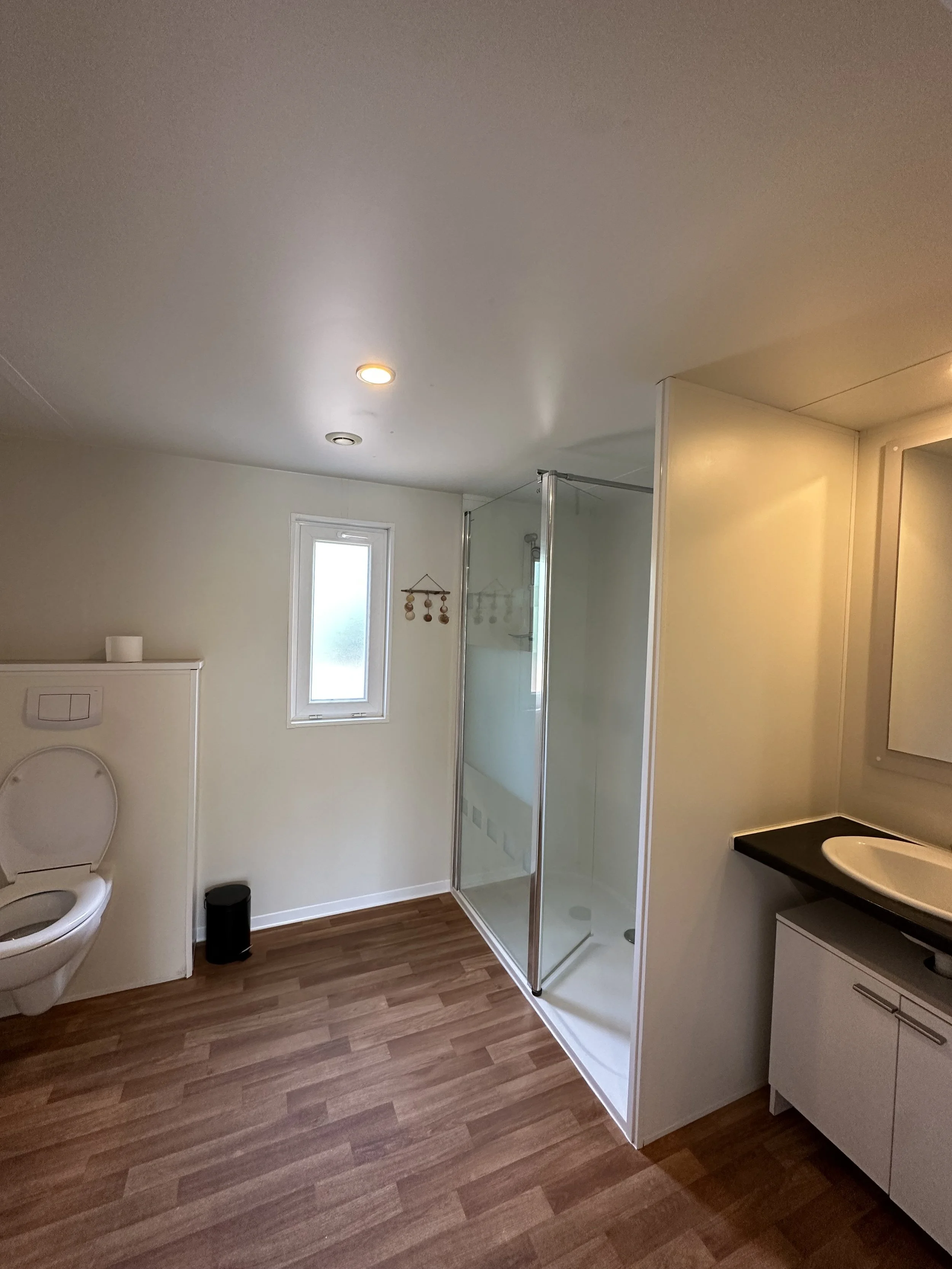 Salle de bain avec toilettes, cabine de douche en verre, évier sur meuble blanc, sol en parquet, petite fenêtre et décoration murale.