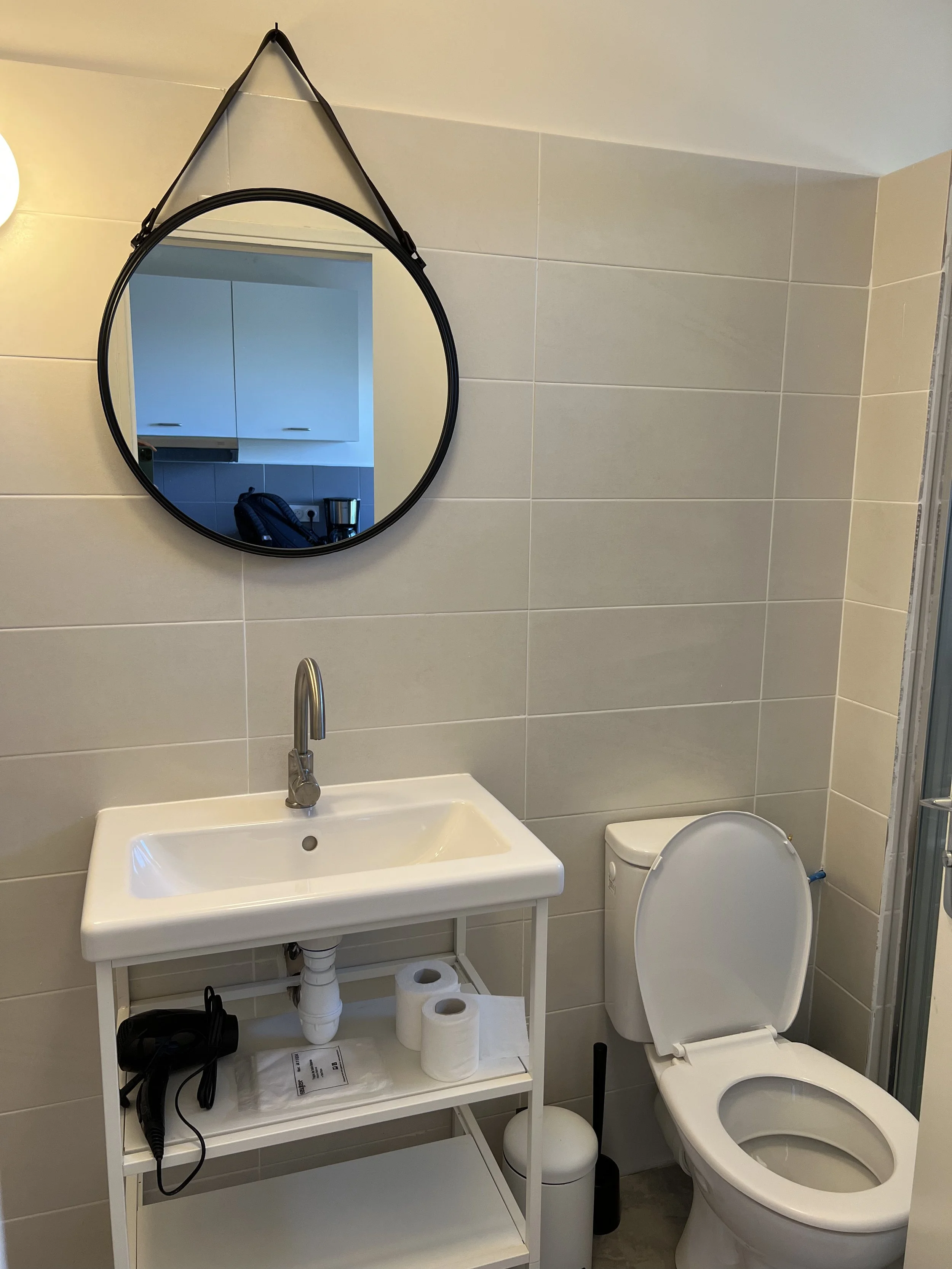 Salle de bain avec un miroir rond suspendu à un mur blanc, un lavabo blanc avec une étagère en dessous contenant un sèche-cheveux, des rouleaux de papier toilette et un sachet, une toilette, une brosse de toilette, un porte-boules, et une porte de douche en verre.