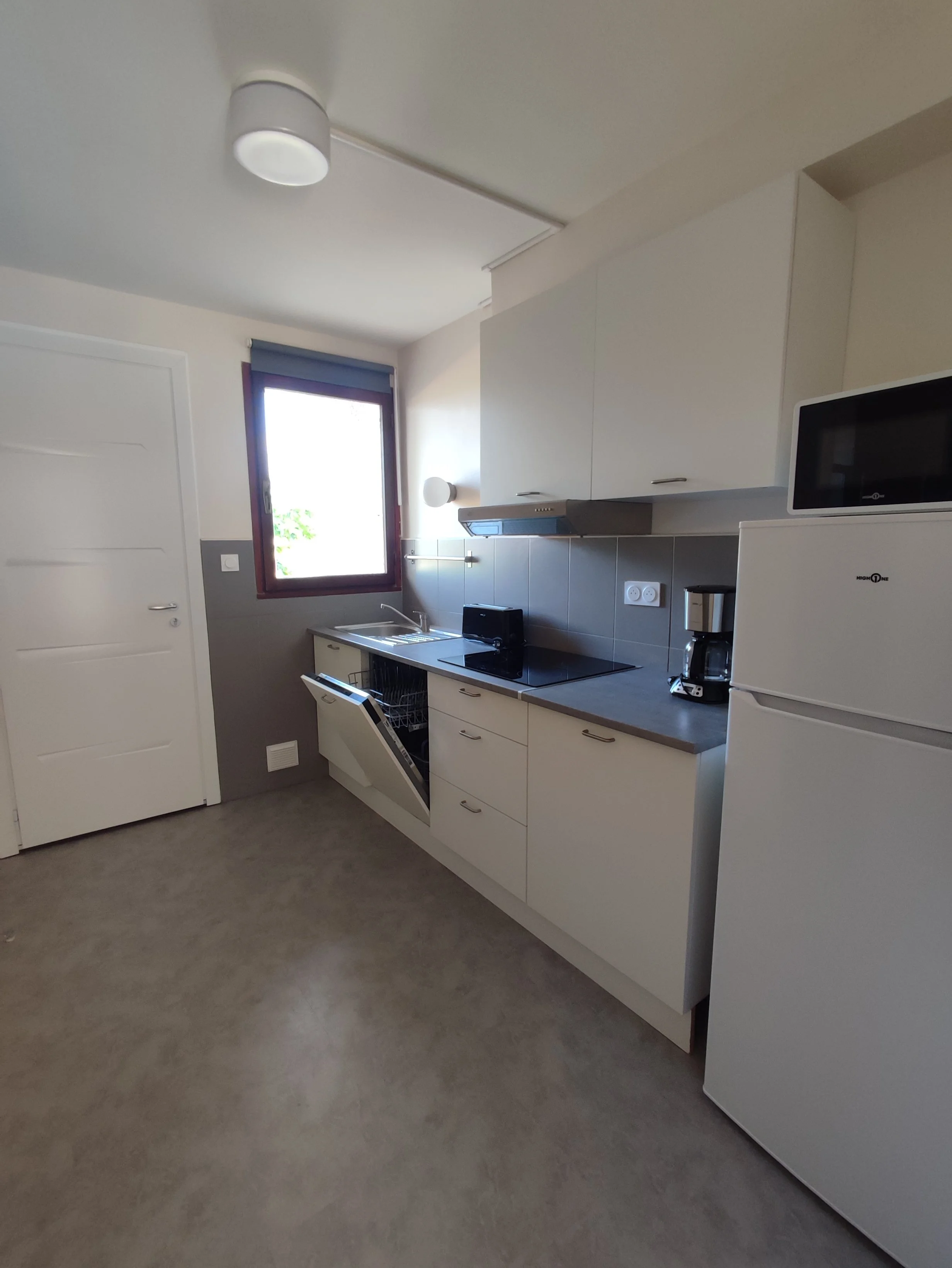 Cuisine moderne avec un évier, un four micro-ondes, un réfrigérateur et divers petits appareils électriques sur le plan de travail, fenêtre et porte blanche.