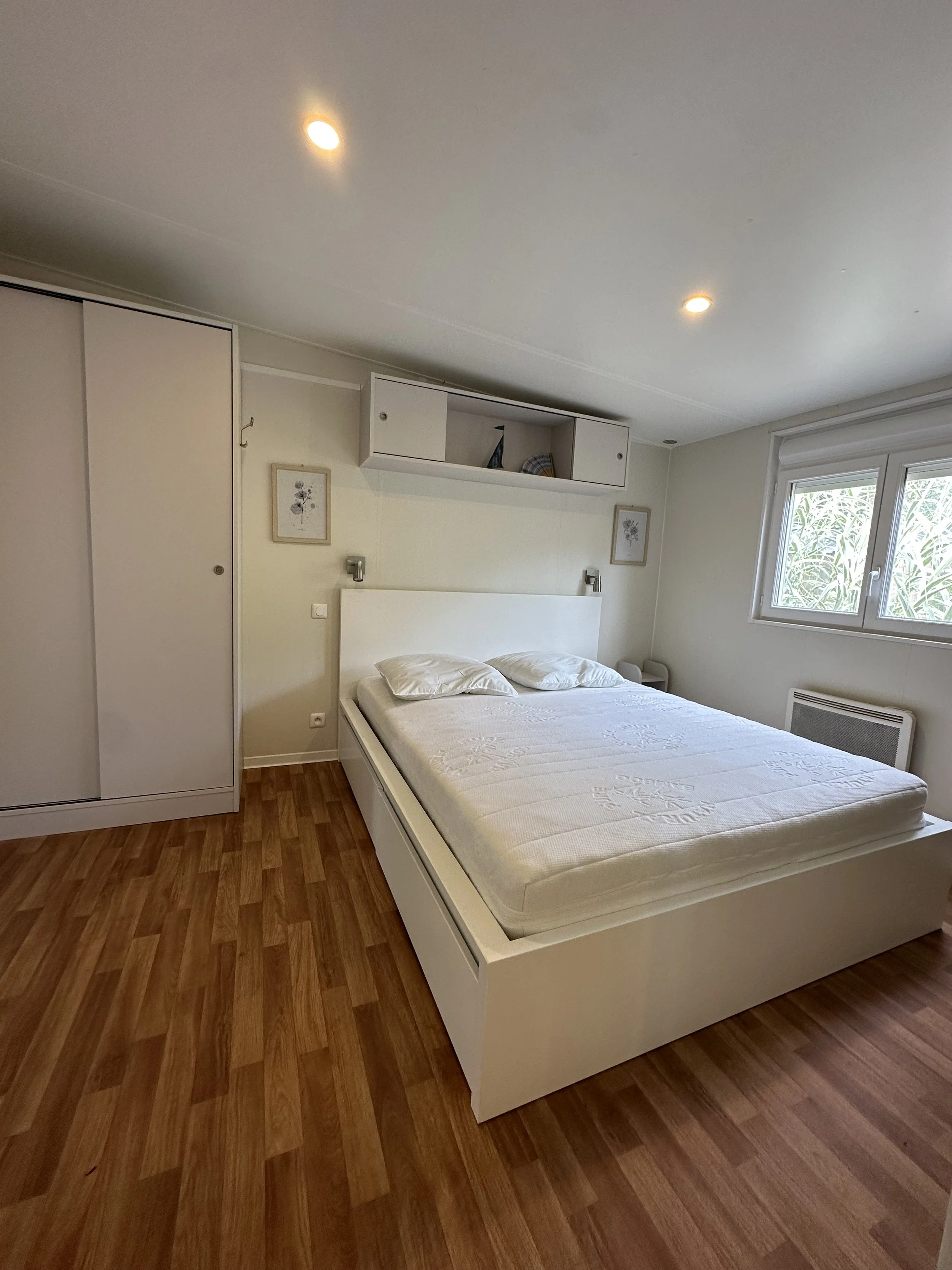 Chambre avec un lit double blanc, deux chevets, une armoire blanche, un mur blanc avec deux images encadrées, une fenêtre laissant entrer de la lumière naturelle, un chauffage électrique en dessous de la fenêtre, et un plafond à deux spots lumineux.