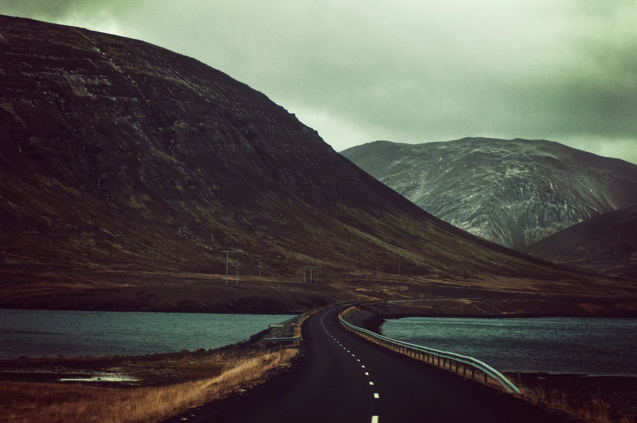 A winding road runs alongside a body of water through a mountainous landscape under a cloudy sky.