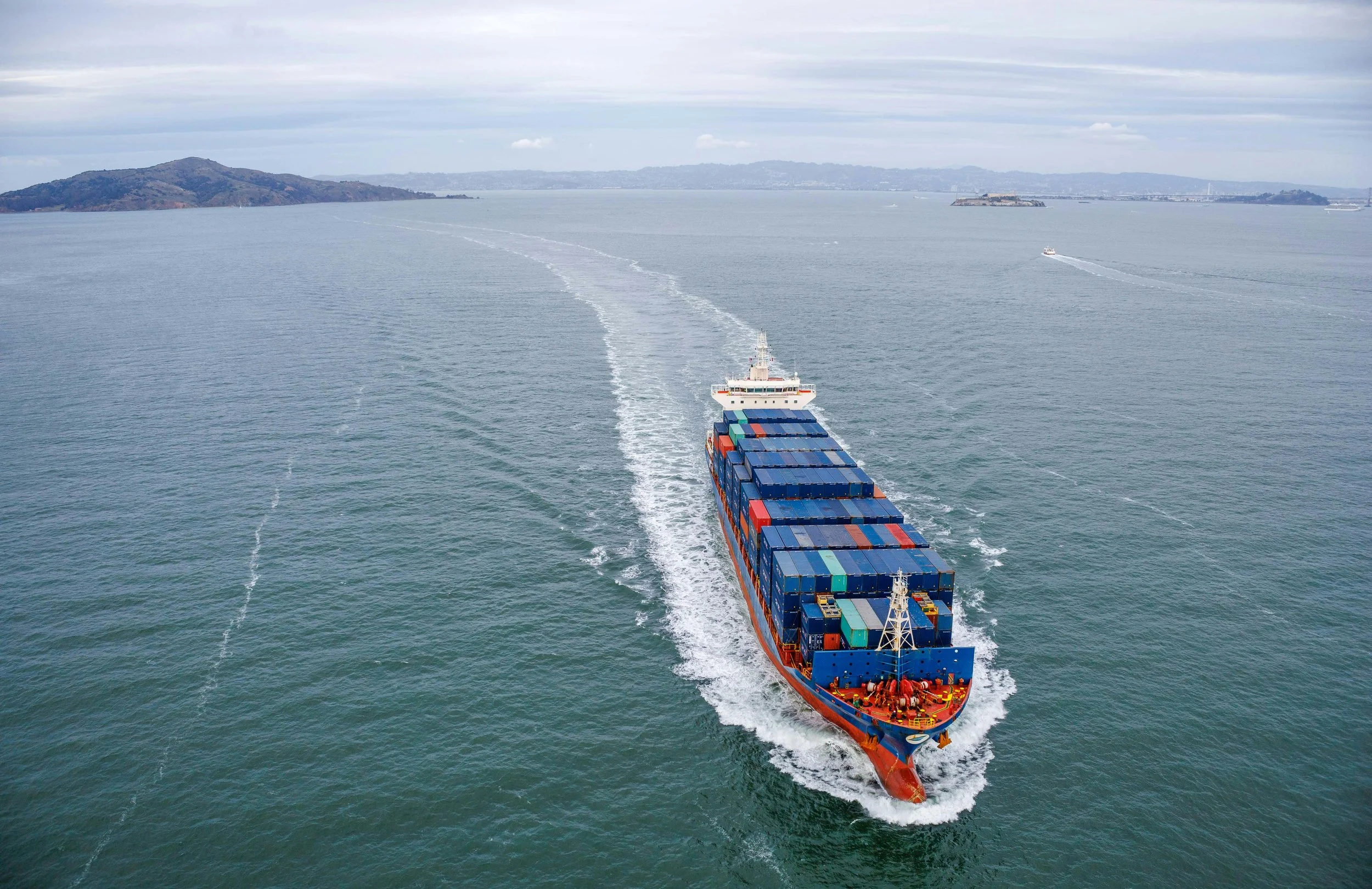 A large cargo ship with numerous shipping containers on deck is sailing in open water, leaving a wake behind. In the distance, there are landmasses and other boats visible under a cloudy sky.
