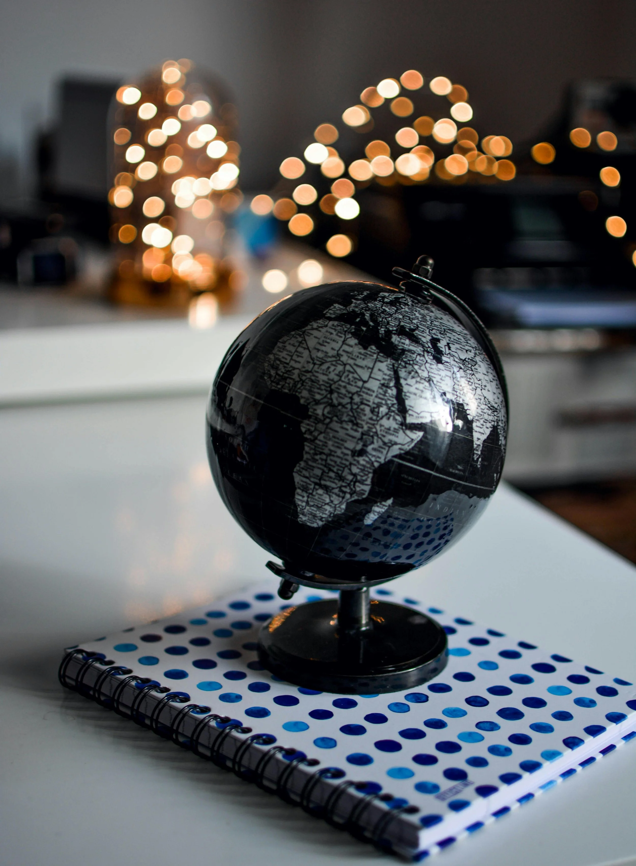 A black globe on a stand resting on a dotted blue and white notebook on a white surface, with blurred warm string lights in the background.