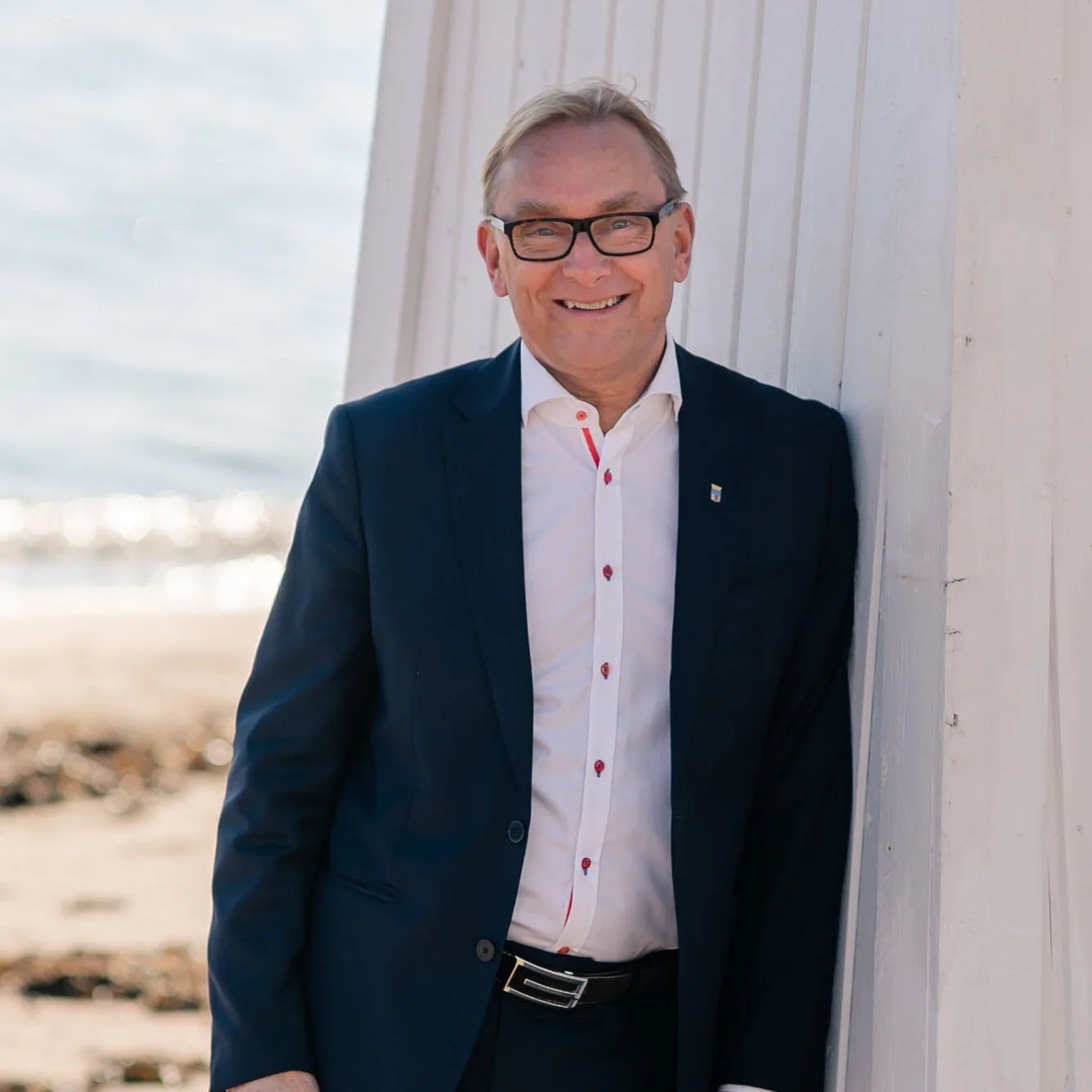 Mies hymyilee ja seisoo rannalla, taustalla meri ja pilvinen taivas.