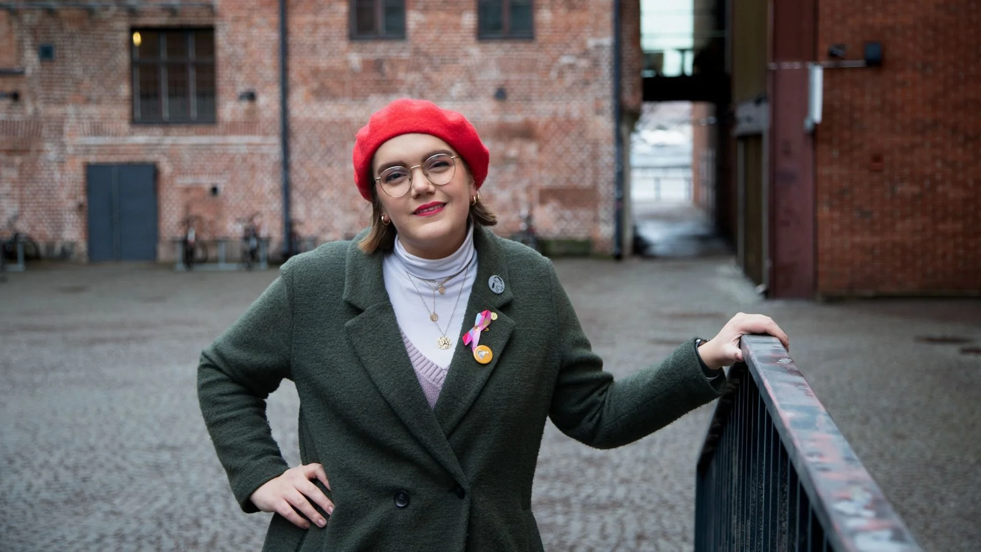 Nainen seisoo ulkona keskeneräisellä betonipihalla, taustalla punatiilinen rakennus, hänellä on harmaa takki, punainen pipo, useita koruja ja muistilappuja, hän hymyilee.