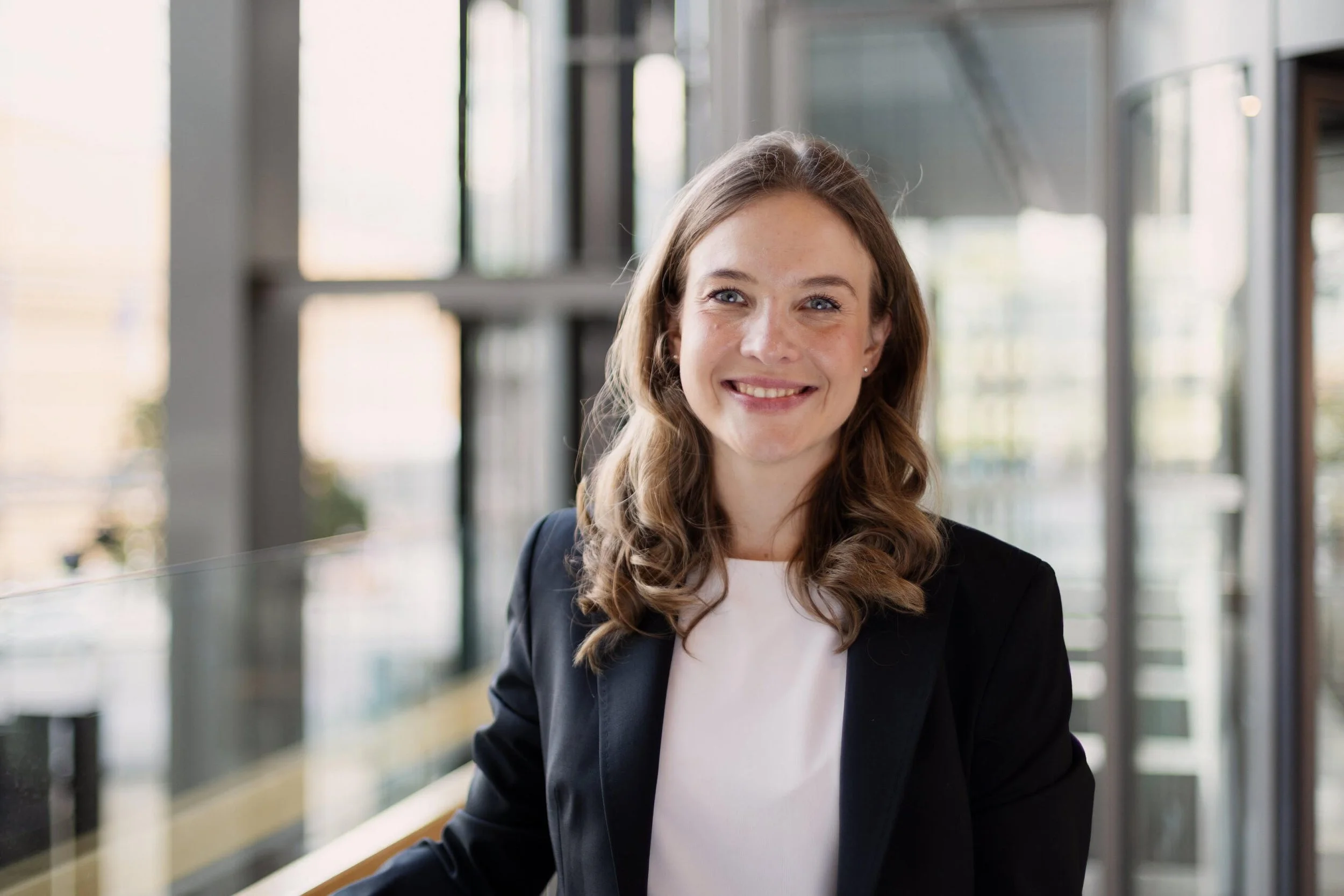 Nuori nainen hymyilee sisätiloissa, taustalla moderni rakennus, iso ikkunoita ja luonnonvaloa. Karin Cederlöf