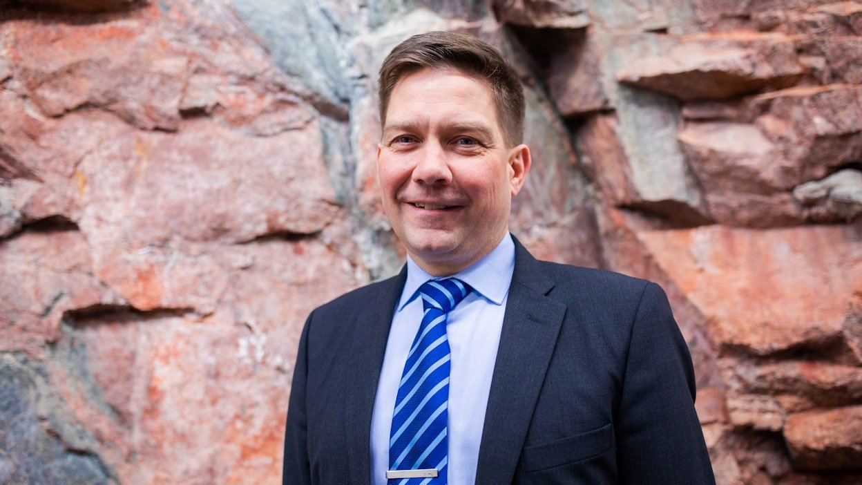 Mies pukeutuu pukuun ja sinivalkoiseen solmioon, taustalla kiviainesseinä.