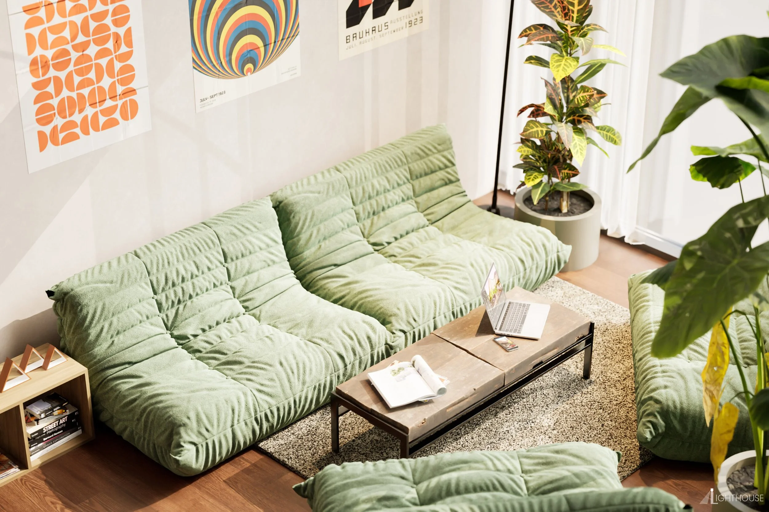A cozy living room with green velvet sofas, a wooden coffee table with a laptop and open magazine, a small bookshelf, a large potted plant, artwork on the wall, and sheer curtains letting in natural light.