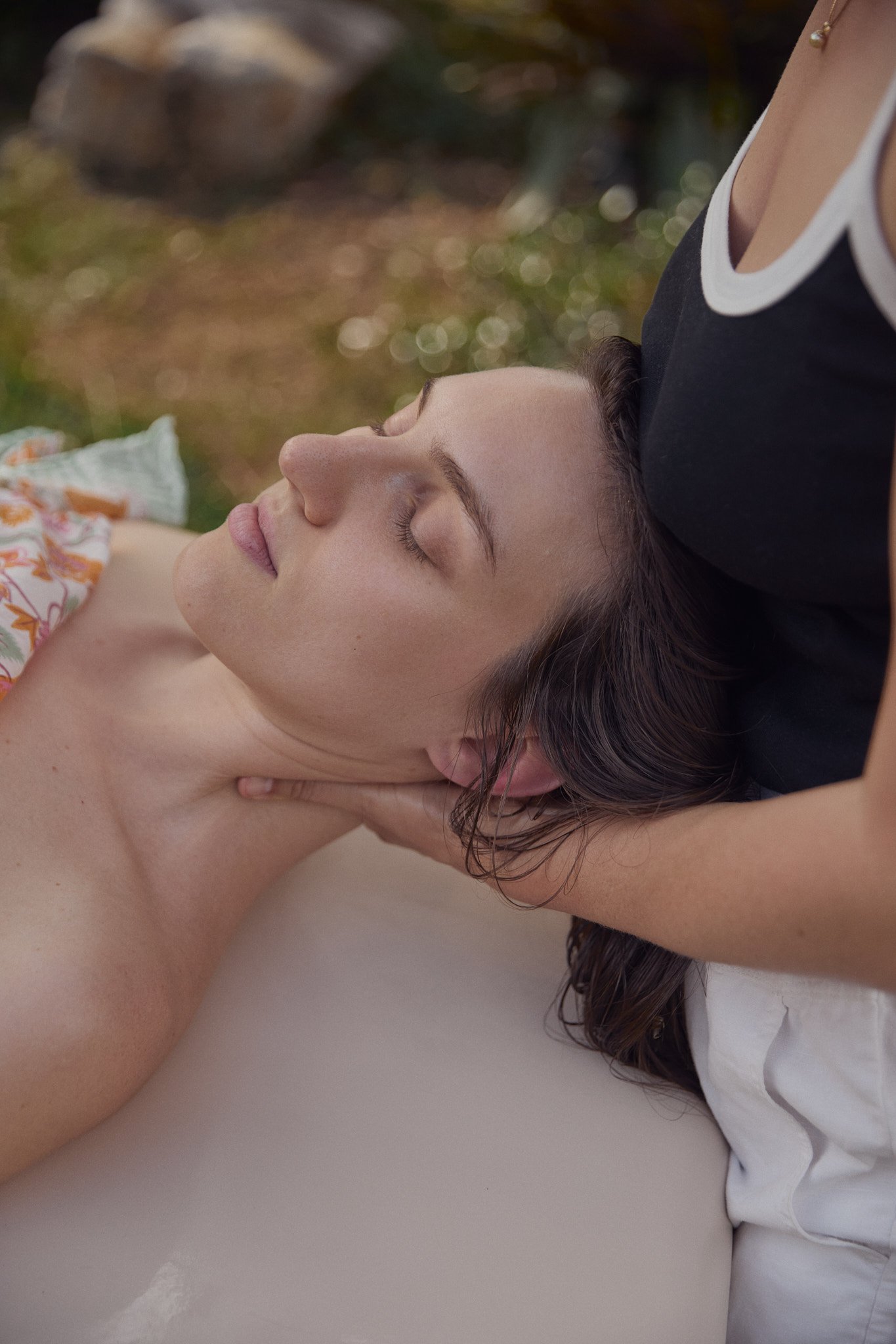 A woman receiving a massage outdoors with her eyes closed, lying on her back, and a masseuse's hands gently supporting her head.