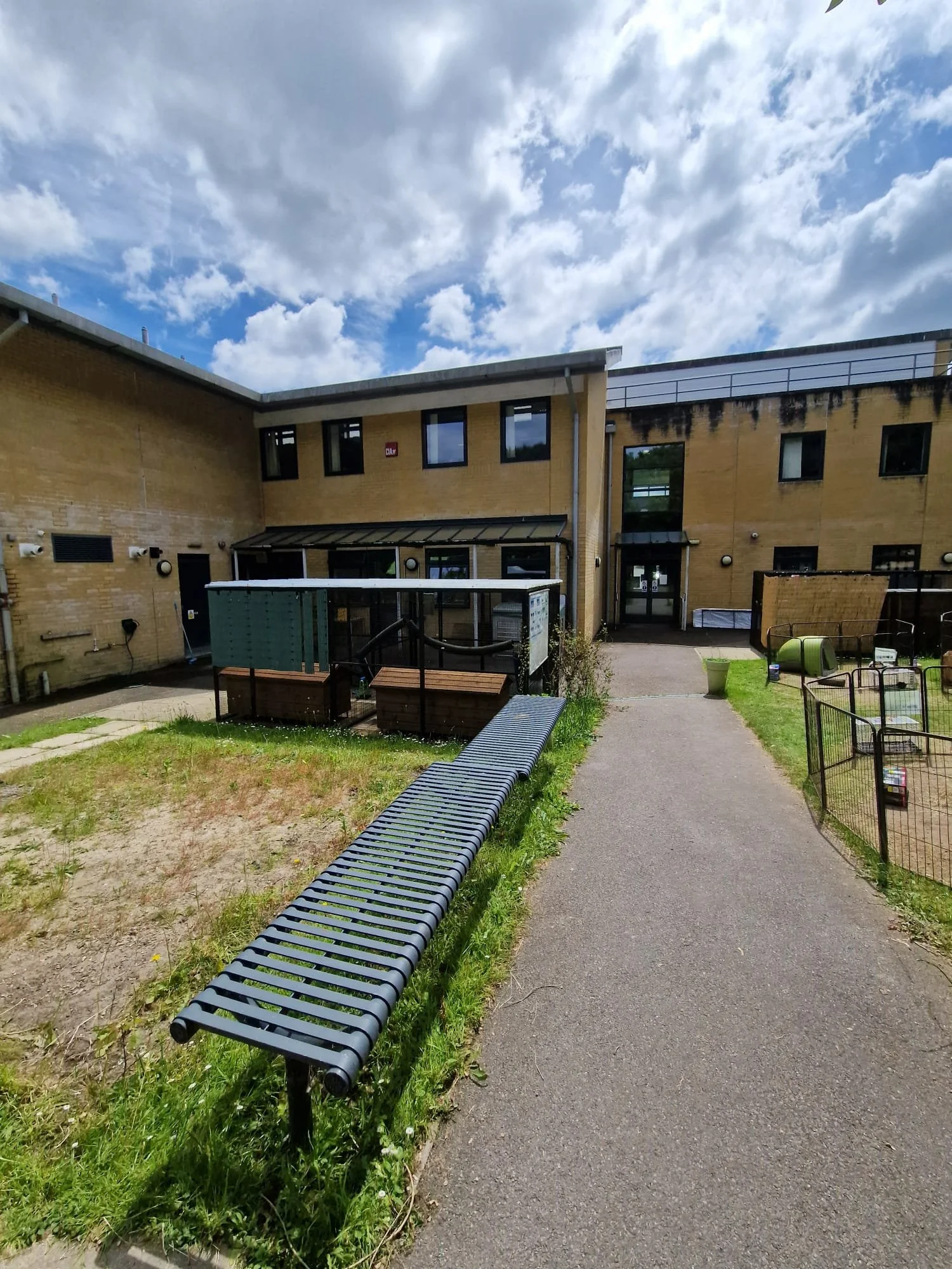 Exterior view of a school or daycare building with a pathway leading to the entrance, a bench along the sidewalk, and small outdoor play areas surrounded by fences under a partly cloudy sky.
