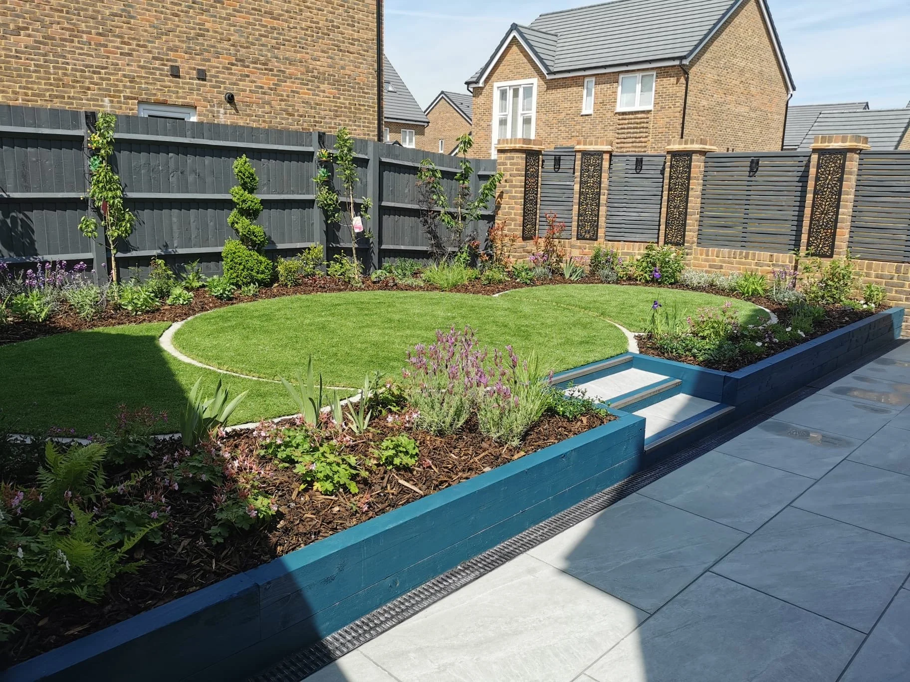 A backyard garden with a circular patch of well-maintained green grass, surrounded by flower beds with various flowers and plants, enclosed by a dark-colored wood fence and brick walls, with white steps leading up to the garden area near a patio with