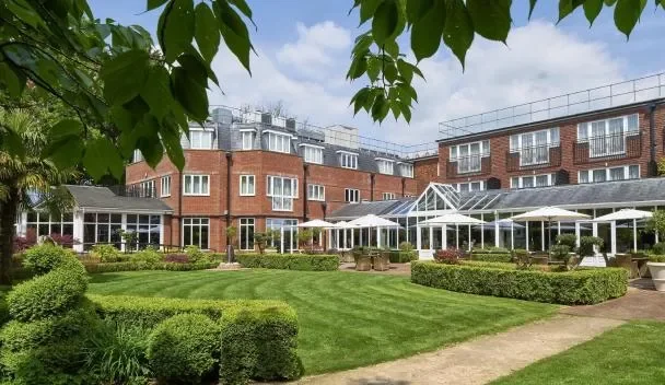 A well-maintained garden with lush green grass, neatly trimmed bushes, and outdoor seating areas with white canopies, with a multi-story brick building in the background under a partly cloudy sky.