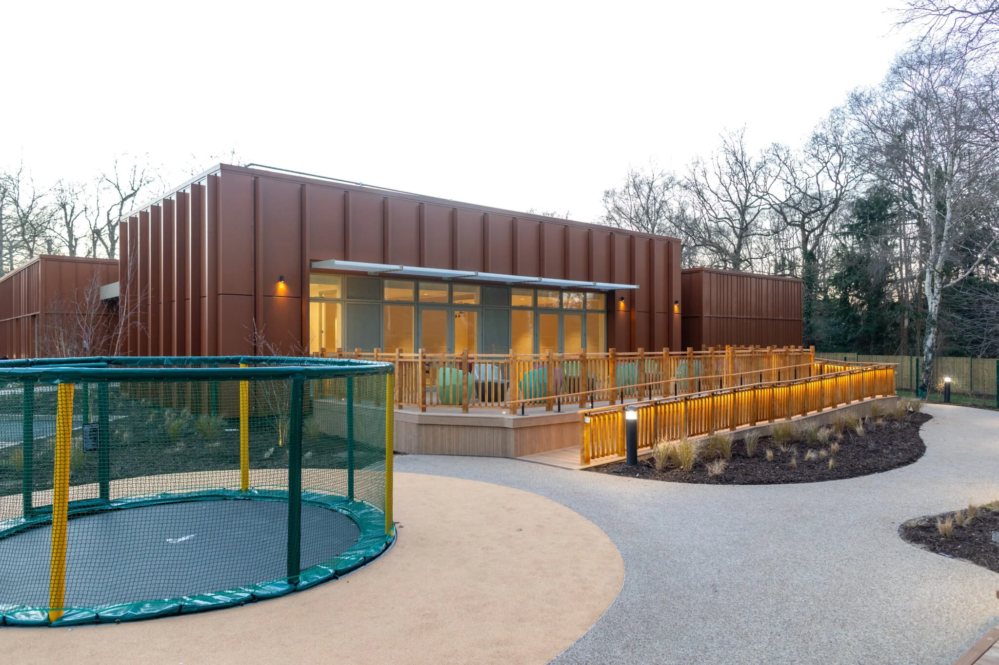 Modern building with a wooden deck and a safety railing, outdoor garden area with a trampoline, paved walking path, and leafless trees in the background.