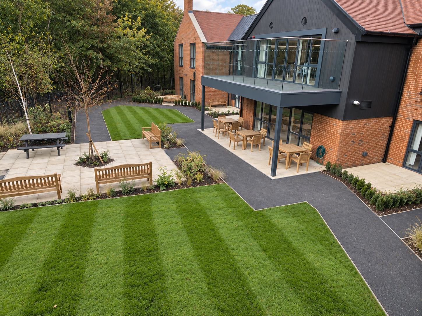 A modern residential backyard with a landscaped lawn, paved walkways, wooden benches, and dining tables with chairs under a balcony, surrounded by trees and shrubs.