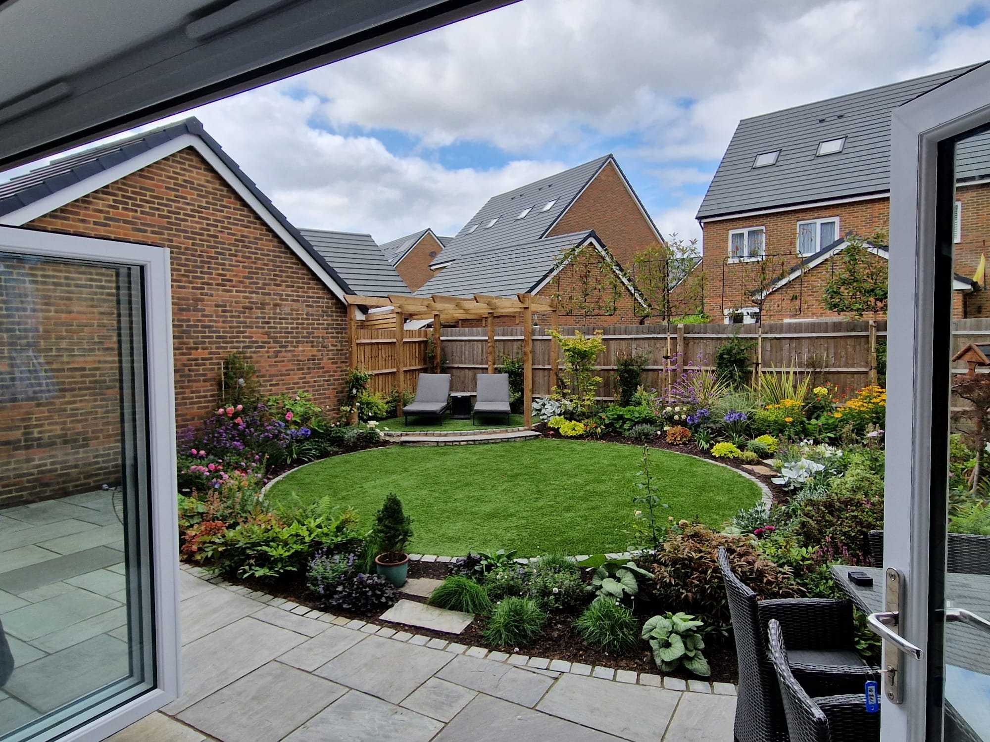 View of a garden through an open patio door, showcasing a well-maintained lawn, colorful flower beds, two lounge chairs with a pergola, and neighboring brick houses with gray roofs.
