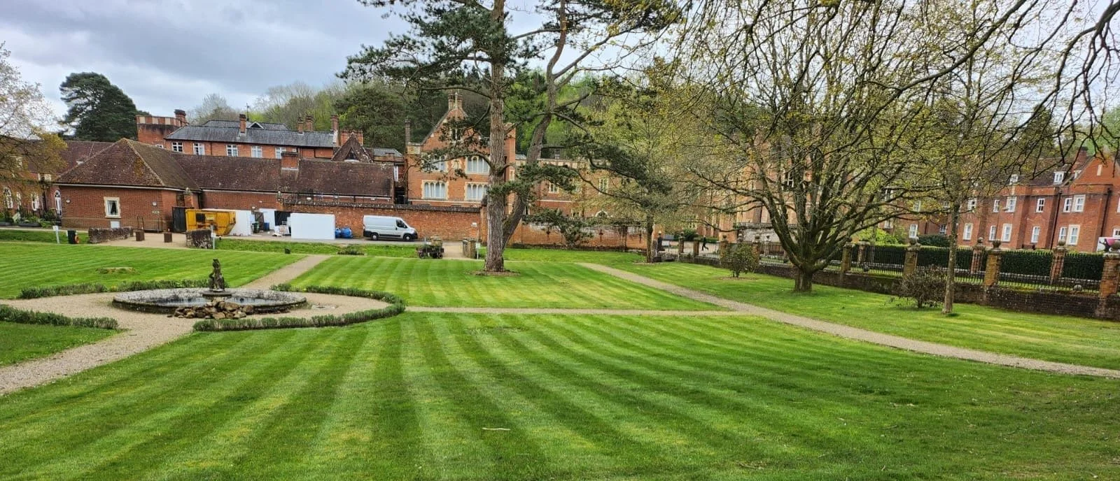 A well-manicured lawn with striped grass, a small fountain, trees, and pathways, set against a backdrop of brick buildings and overcast sky.