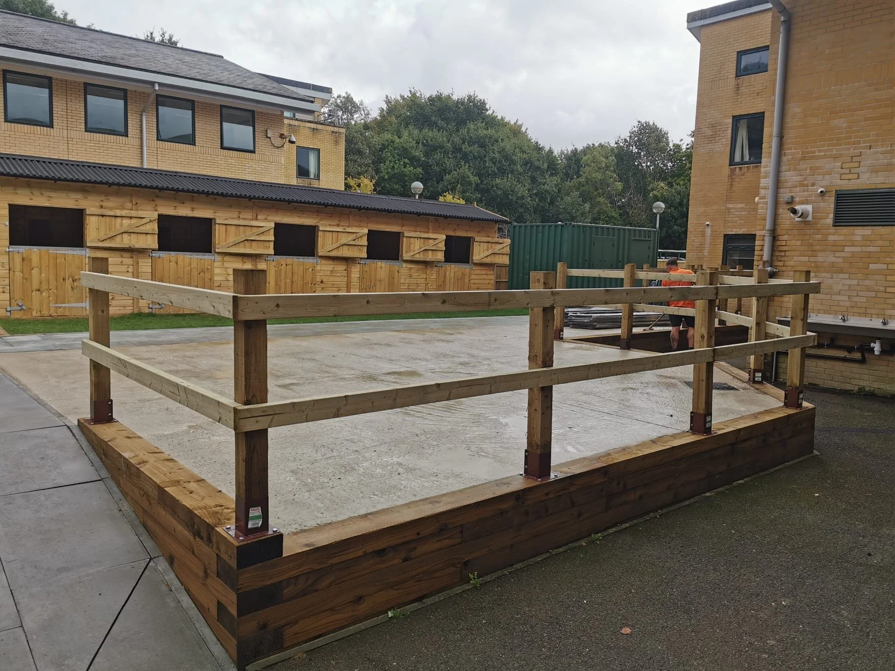 A wooden deck with a railing under construction in a parking lot outside residential buildings, with a person working nearby.