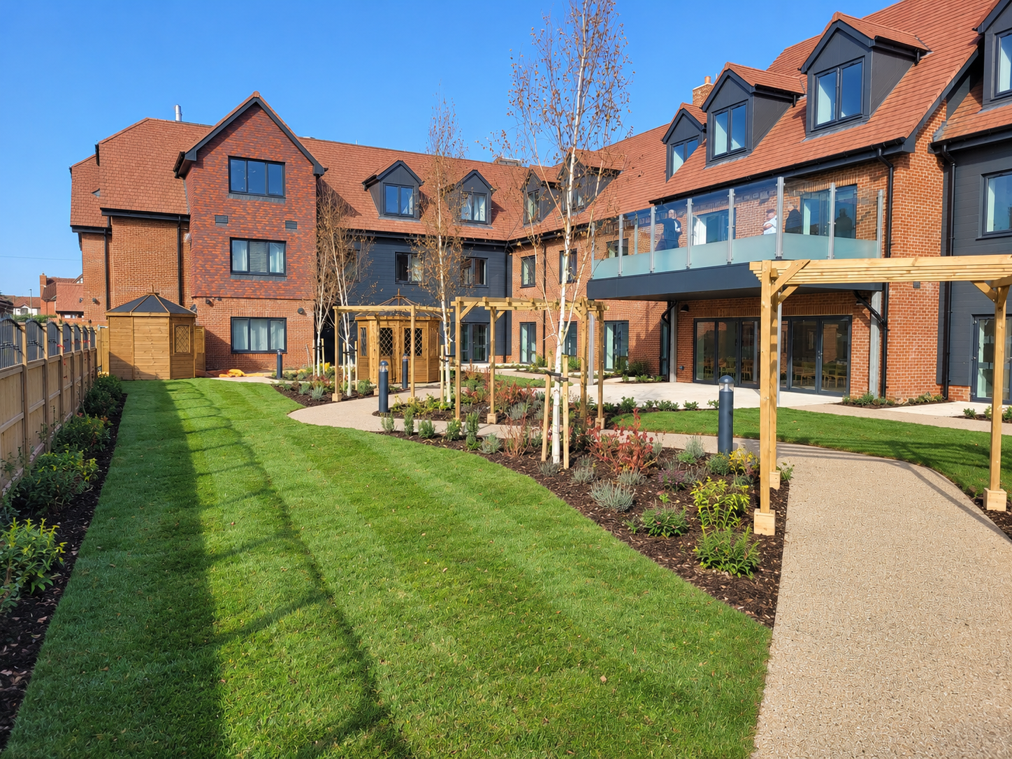 Modern residential apartment building with red brick and black accents, glass balconies, and a landscaped courtyard with grass, young trees, pathways, and garden beds.
