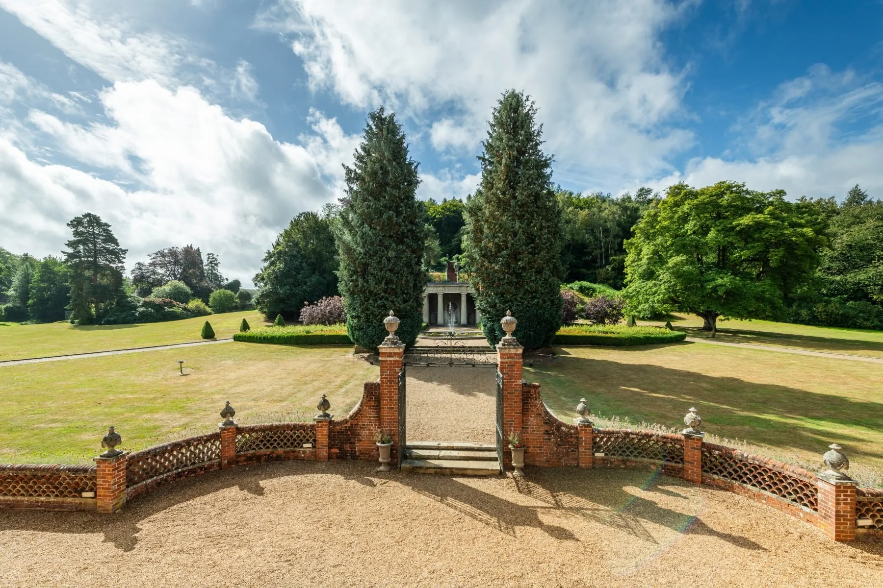 View of a formal garden with a central fountain, surrounded by tall trees, green grass, and a bright blue sky with white clouds.
